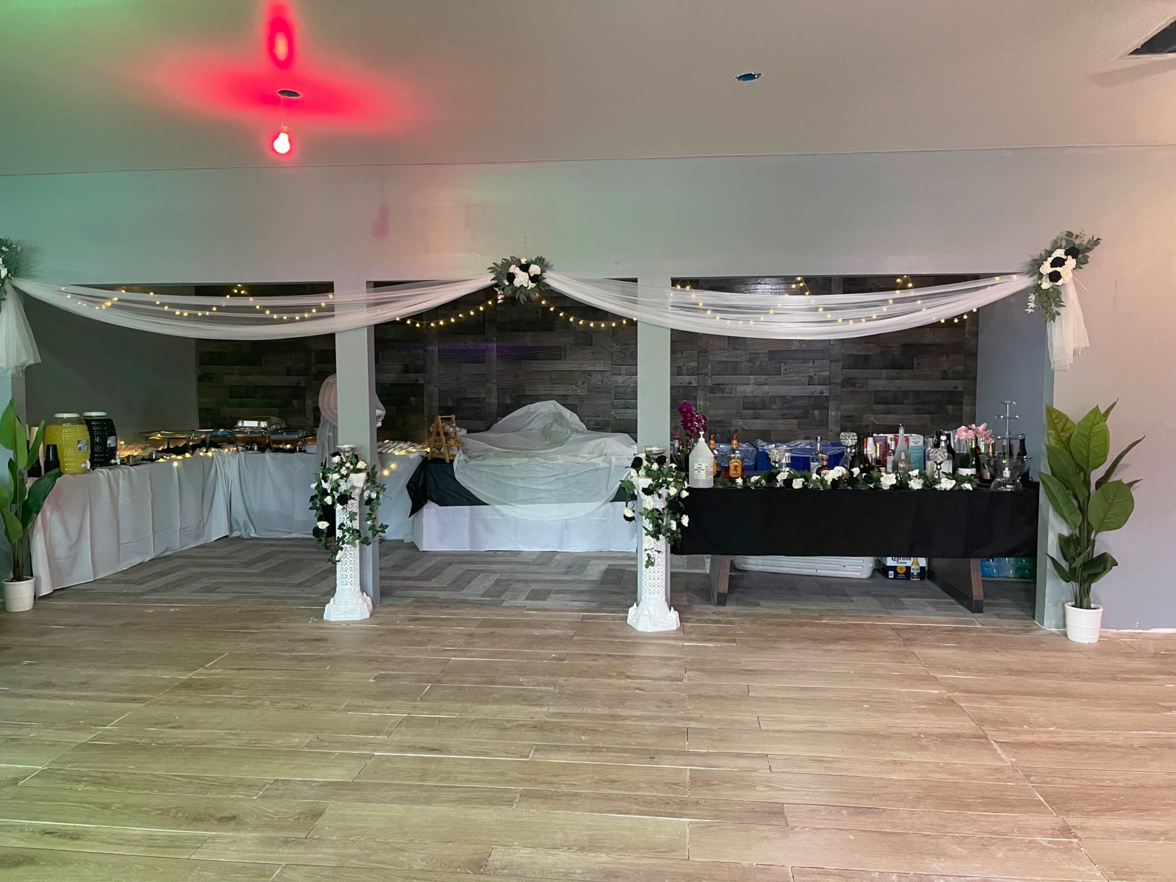 The image features a decorated indoor venue with tables set for an event, displaying food and beverages, adorned with white drapes and fairy lights.