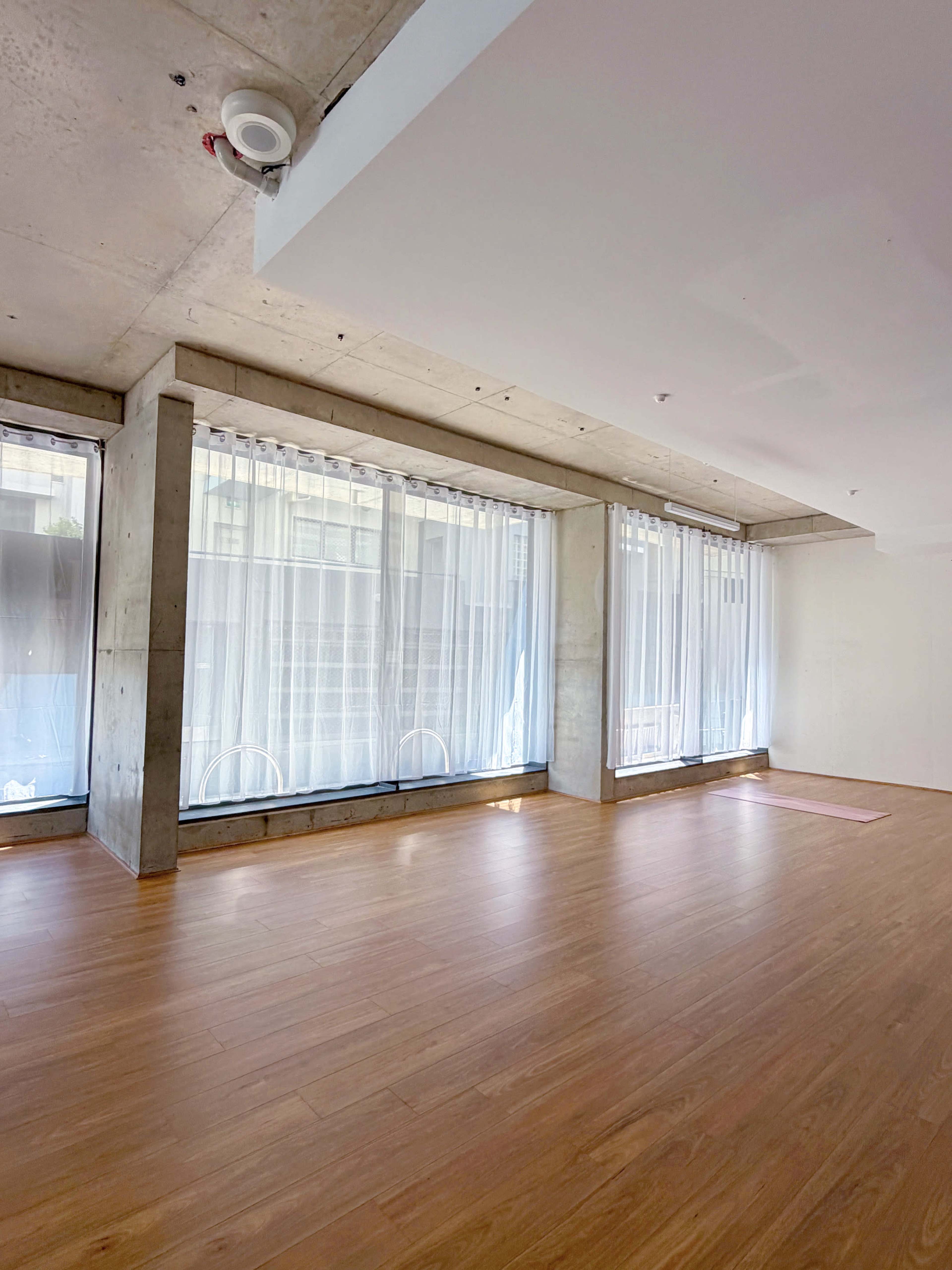 The image shows an empty room with large windows draped in sheer curtains, featuring a wooden floor and concrete walls.