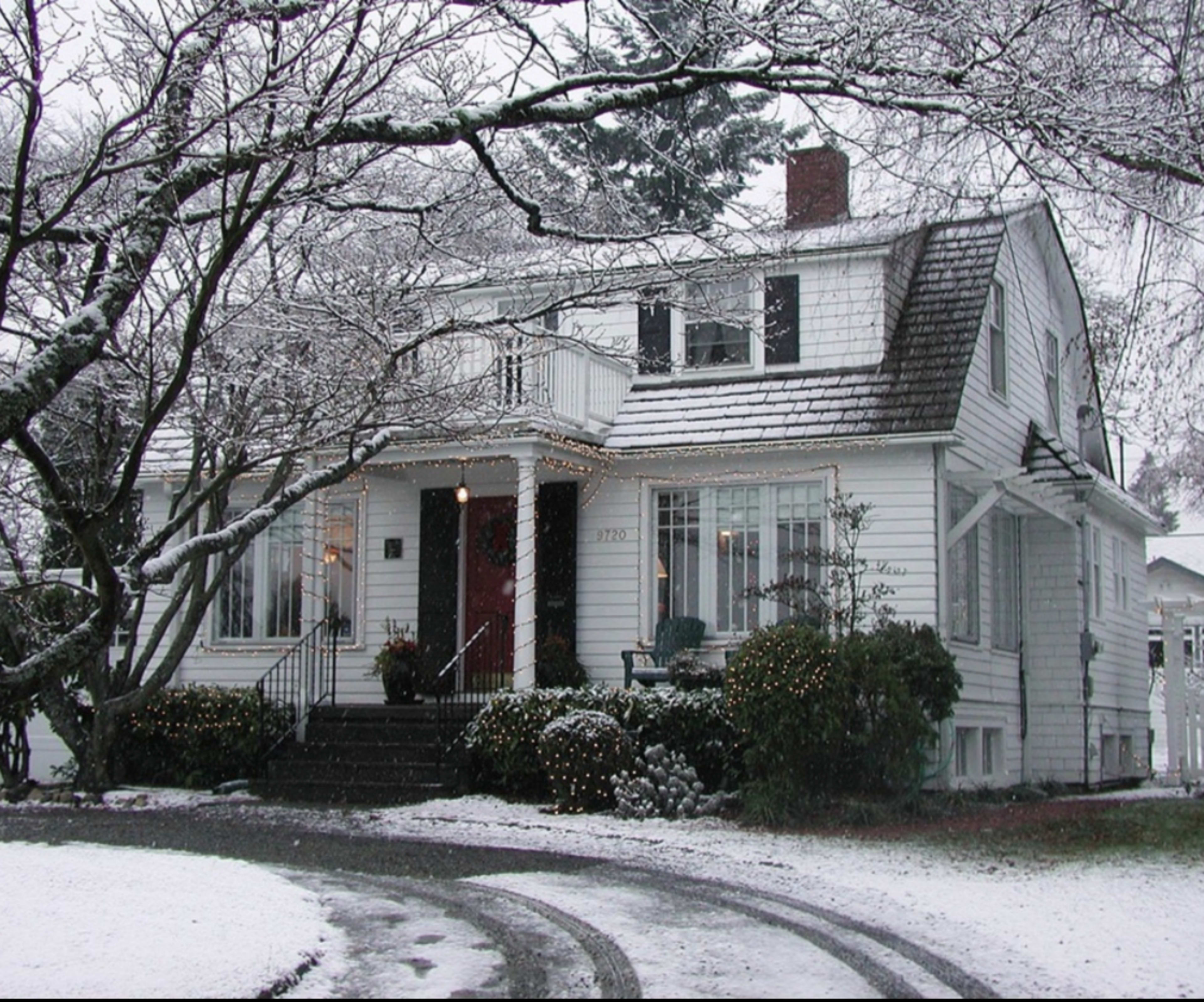 A white two-story house is covered in snow, decorated with lights and surrounded by trees.