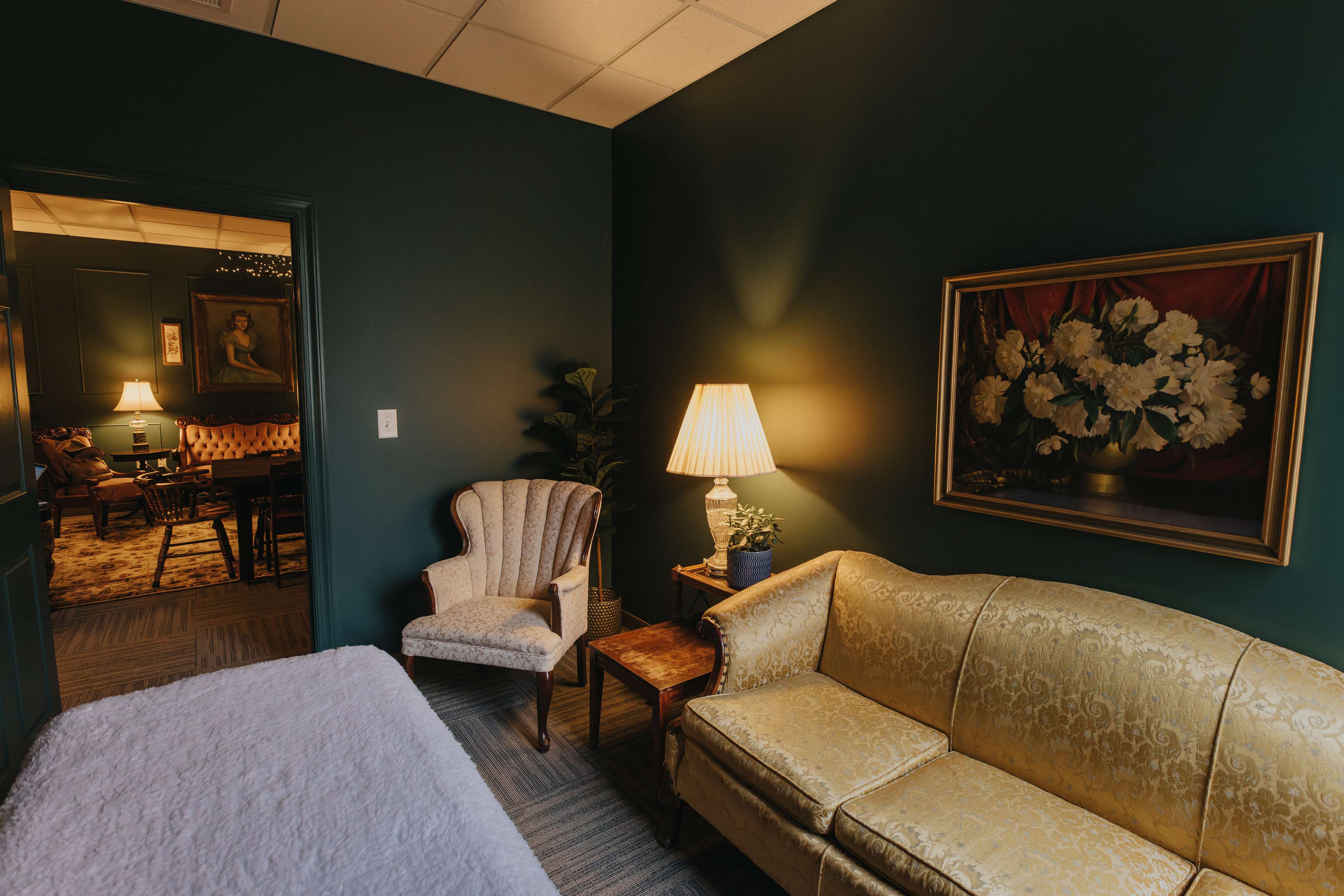 A small room with a gold-colored couch, a patterned armchair, a lamp, and a floral painting on the wall.