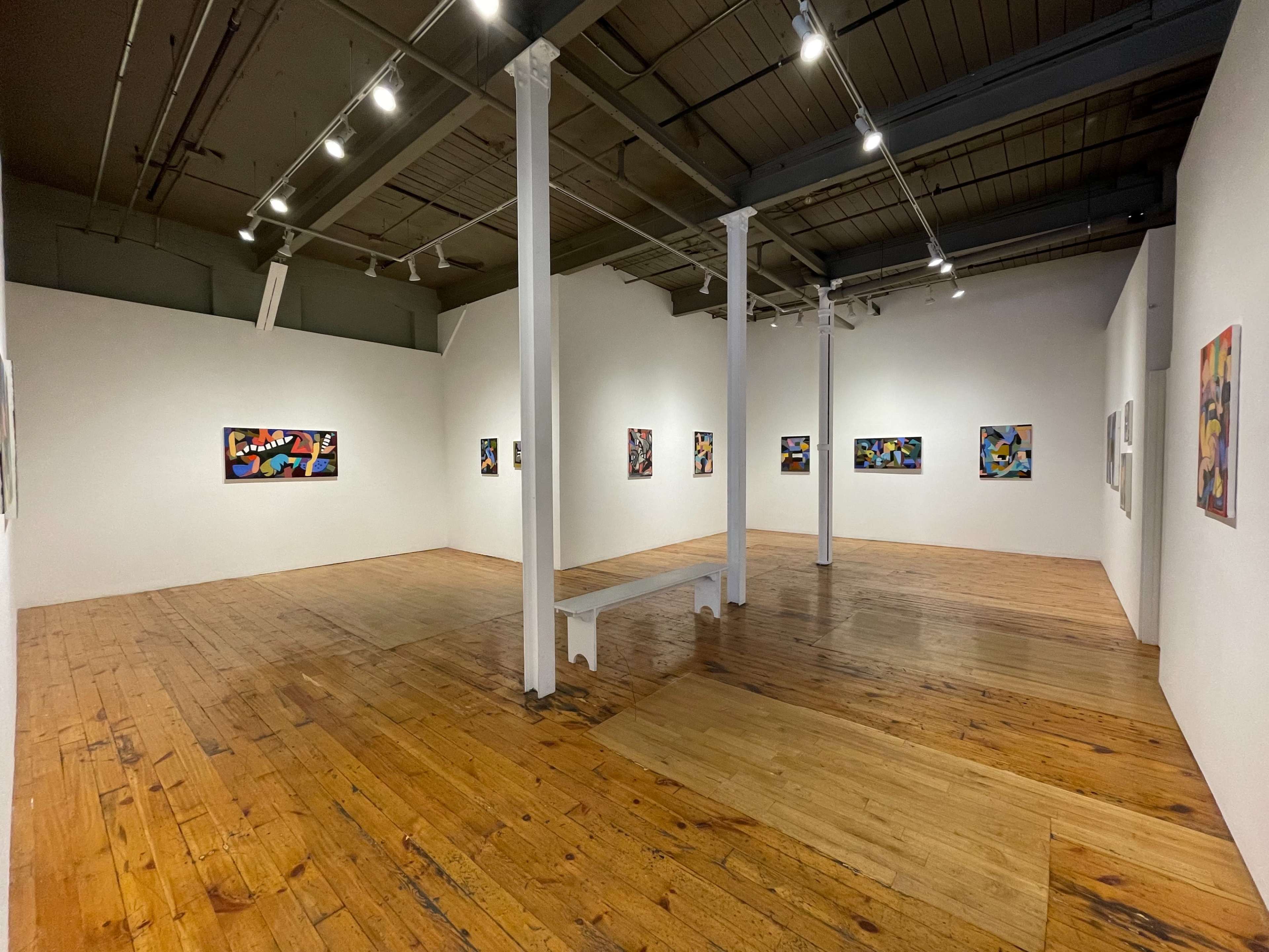 A spacious art gallery with white walls, wooden flooring, and several colorful paintings displayed along the walls.