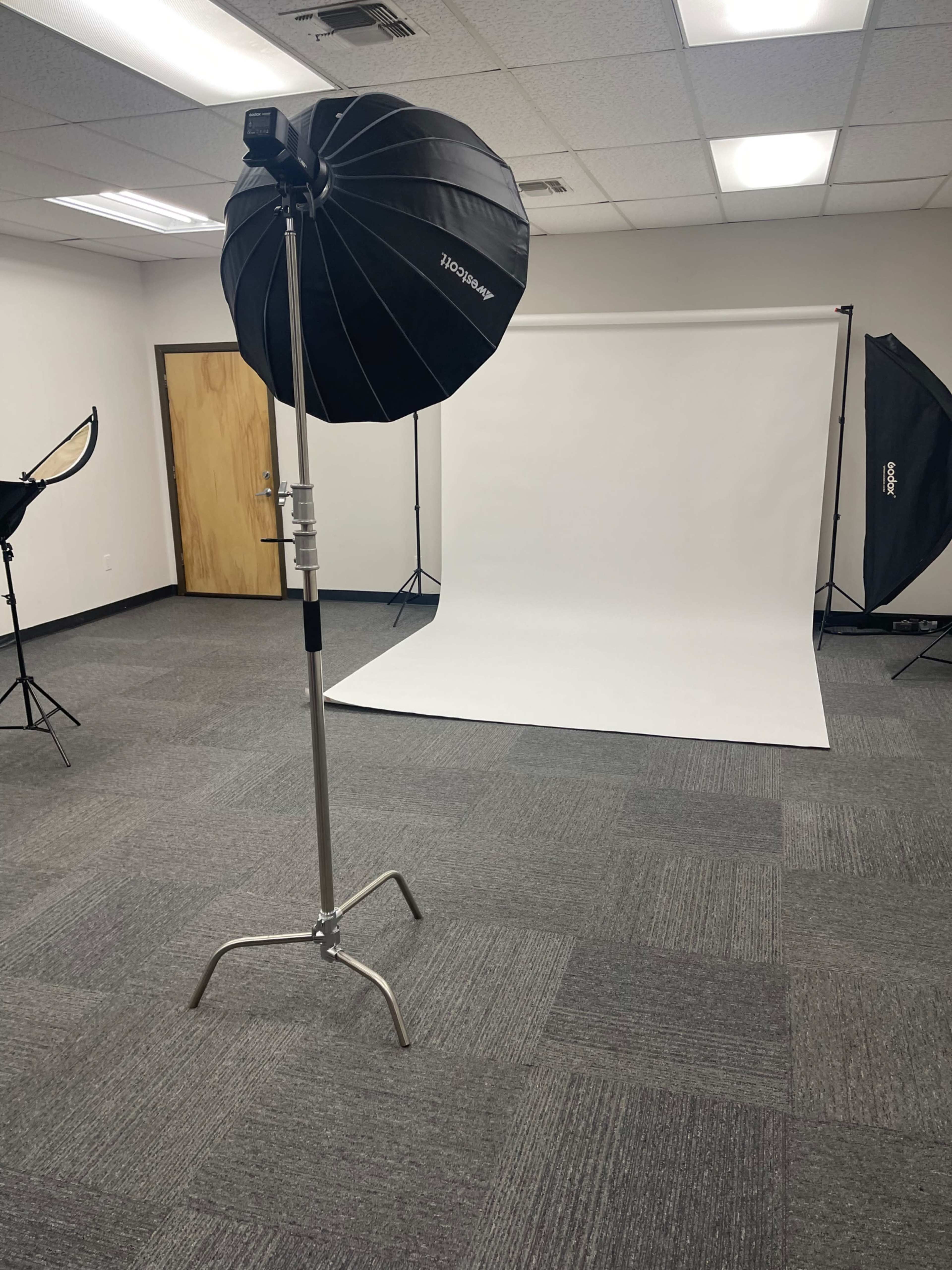 The image shows a photography studio setup with a large softbox light, a backdrop stand with a white backdrop, and various lighting equipment arranged in the room.