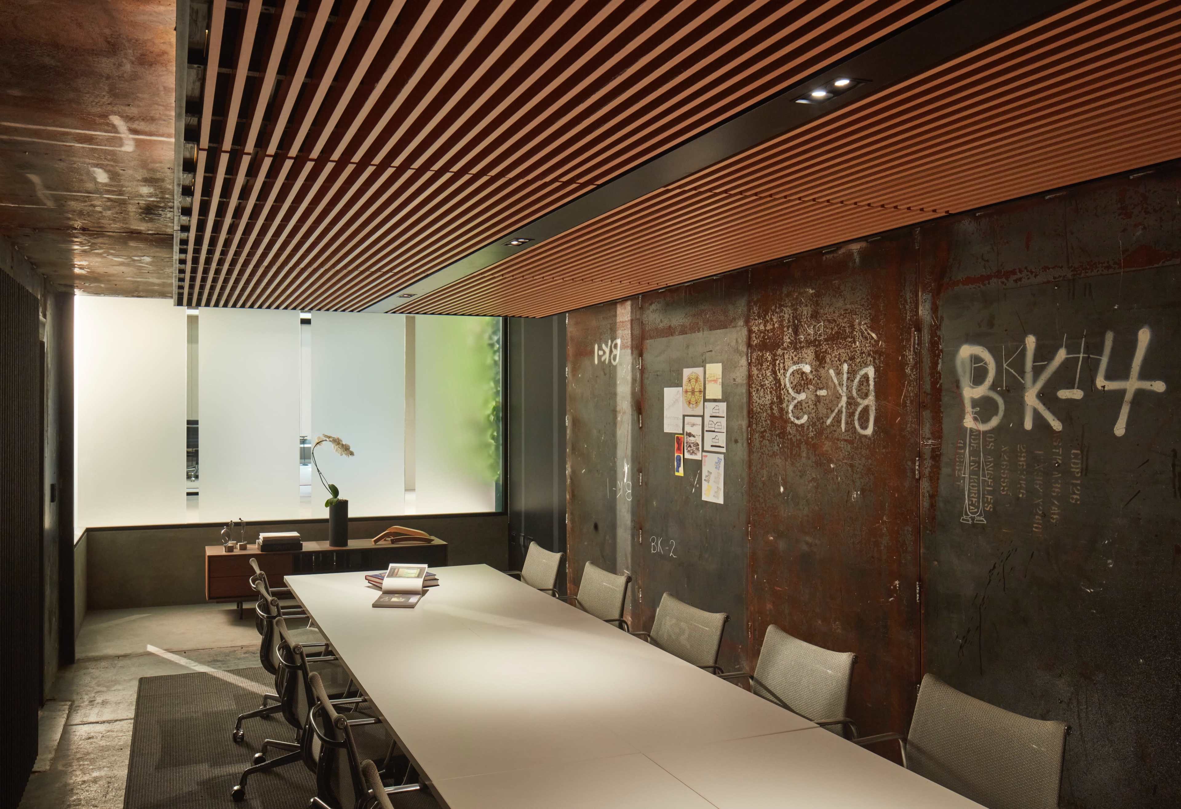 A modern conference room with a long white table, metal walls, and wooden ceiling slats, accented by minimal decor and a large window providing natural light.