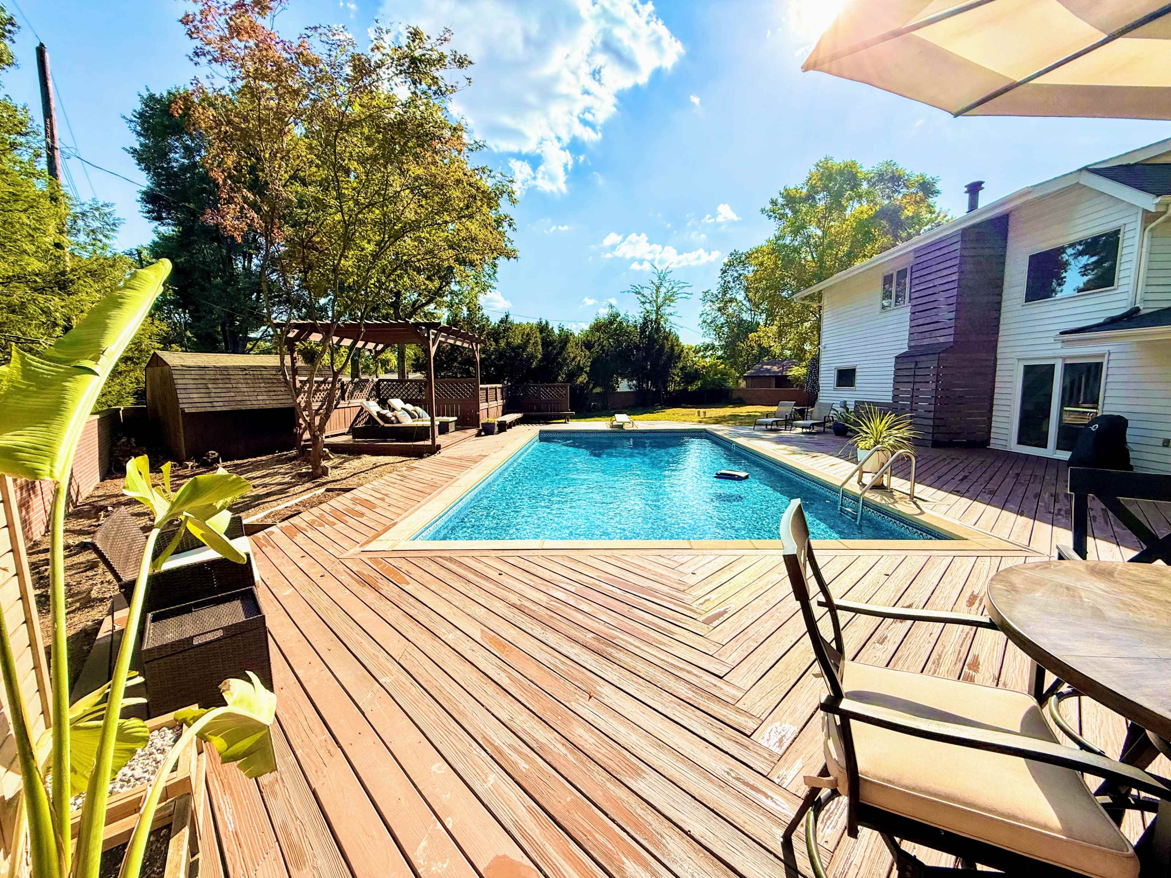 The image shows a backyard with a swimming pool surrounded by a wooden deck, with lounge chairs and a dining area nearby.