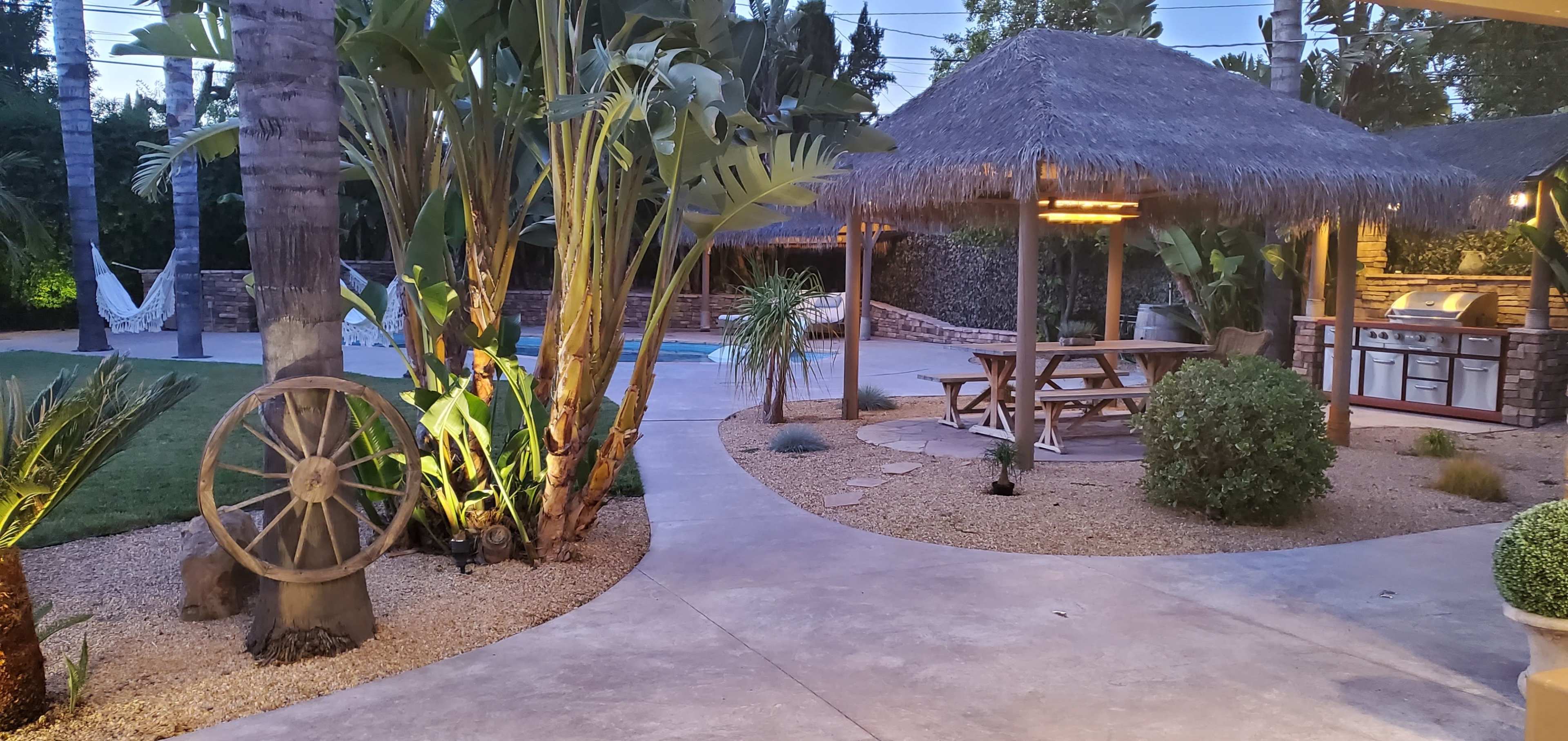 A backyard with a swimming pool, a thatched-roof gazebo with picnic seating, and tropical plants surrounding a stone pathway.