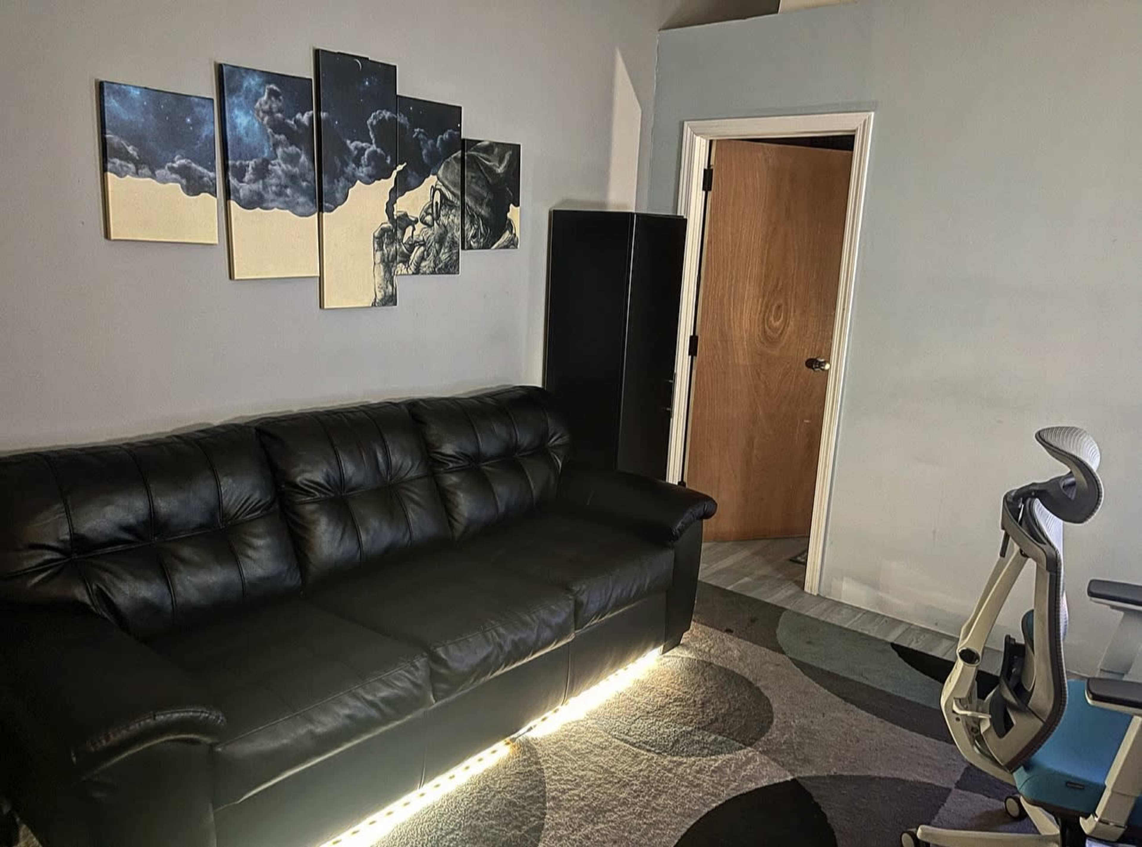 A black sofa with low lighting underneath, positioned in front of a wall featuring abstract art, with a door and a dark cabinet visible in the background.