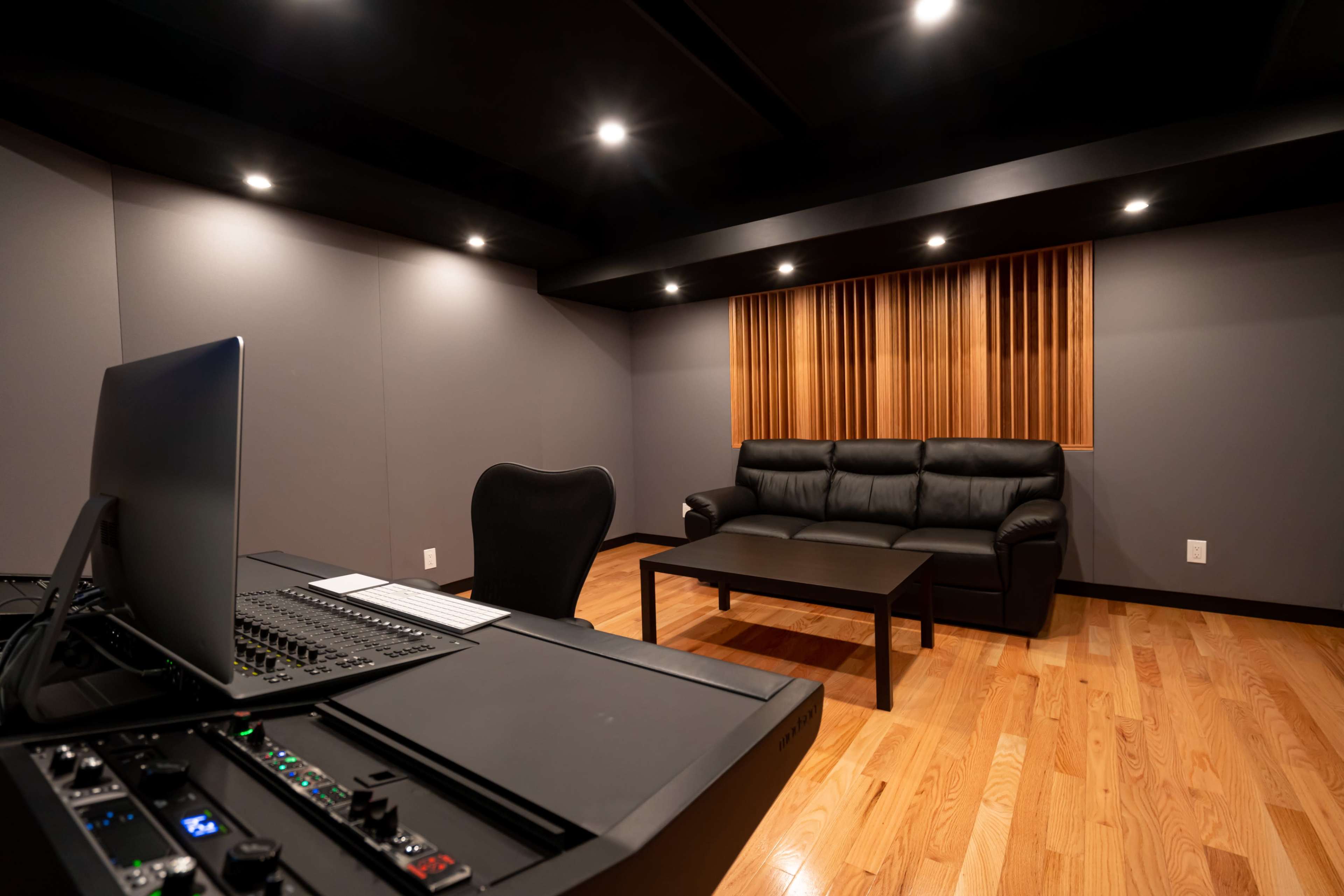 A modern control room with a black desk equipped with audio mixing equipment, a black chair, and a leather couch positioned near a wooden coffee table against a wall with soundproofing panels.