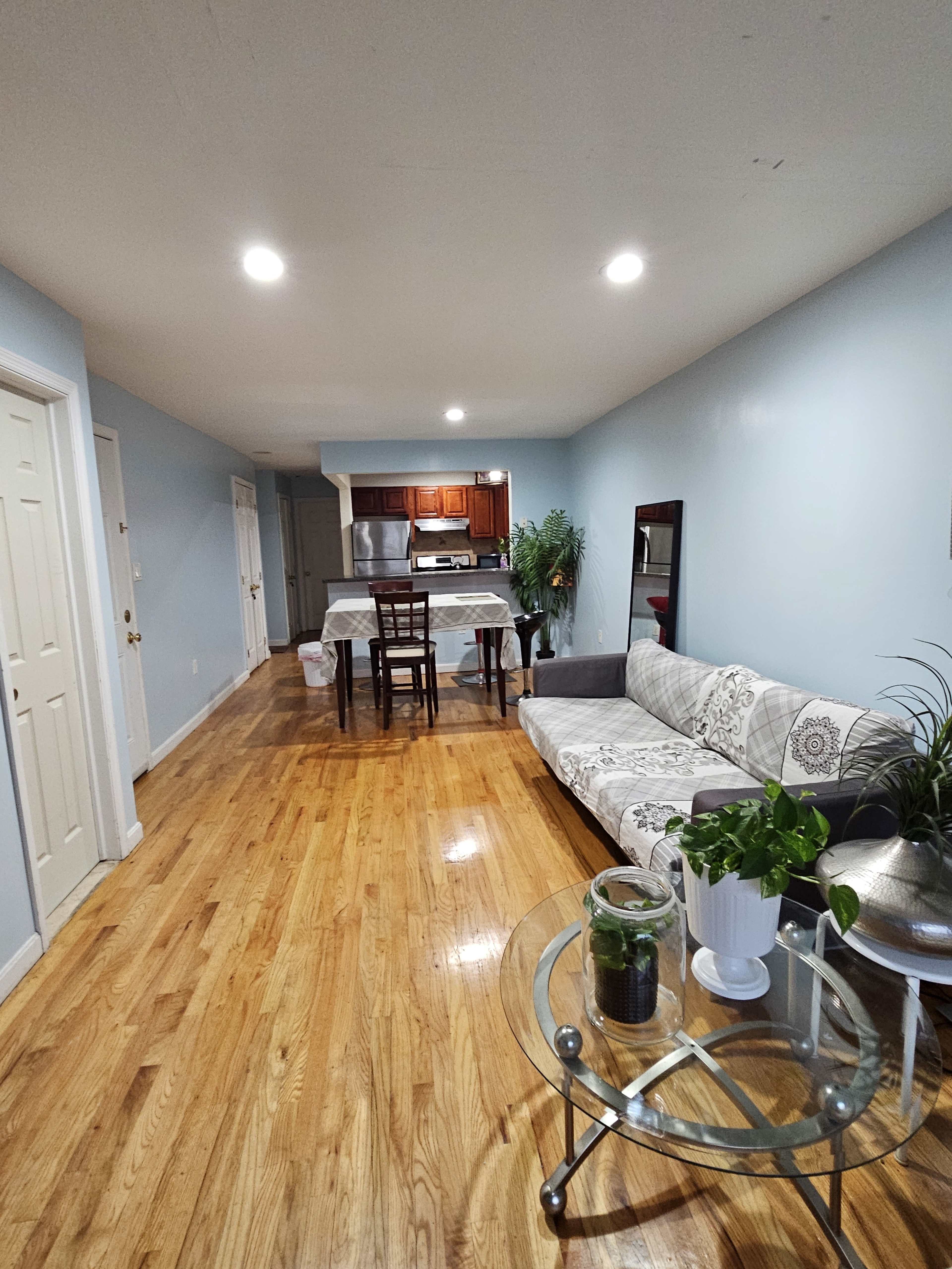 A living area with a sofa, a dining table with chairs, and a kitchen area in the background, all featuring hardwood flooring and light blue walls.