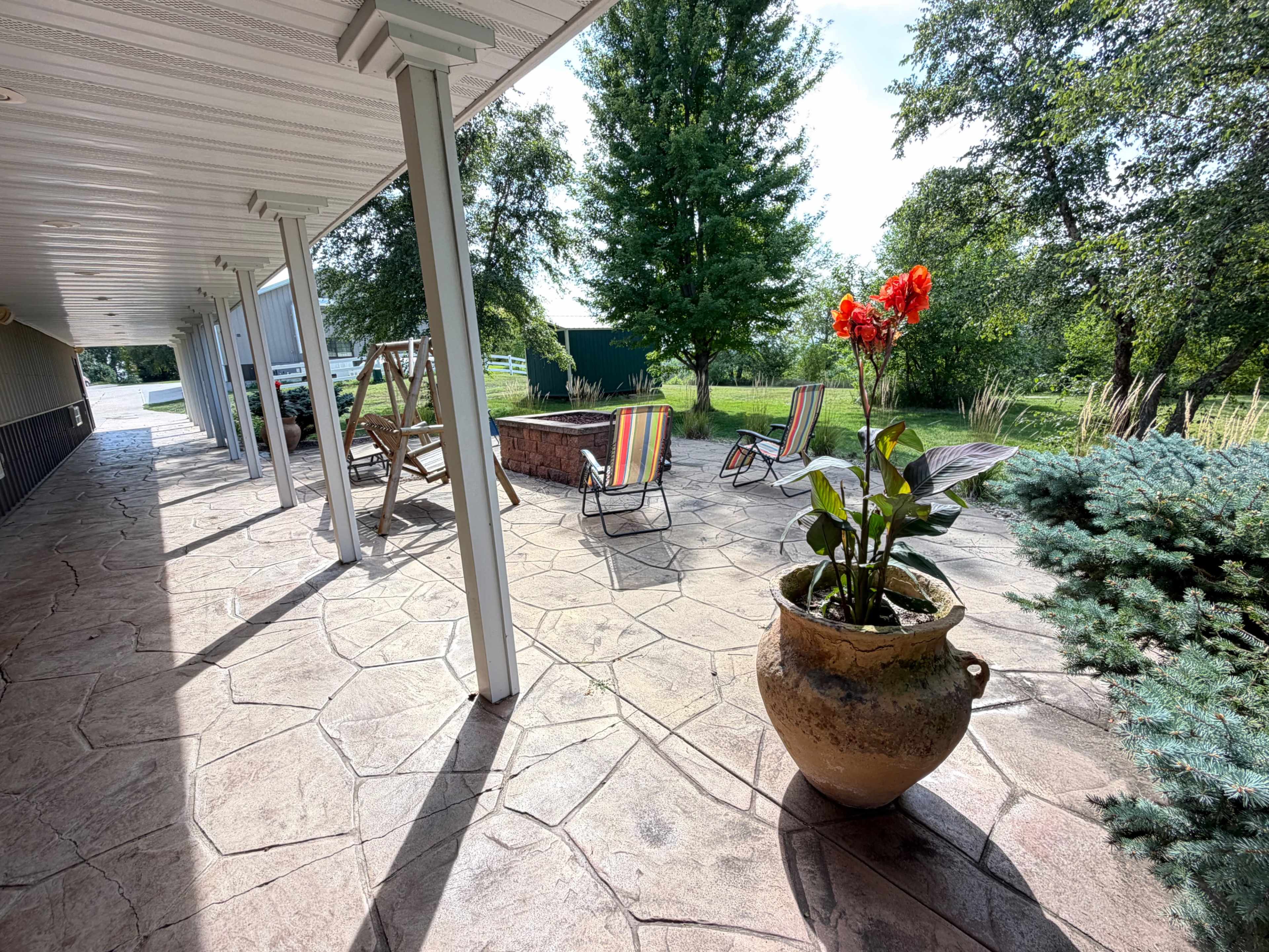 The image shows a covered patio area with lounge chairs and a fire pit, surrounded by greenery and decorative plants.