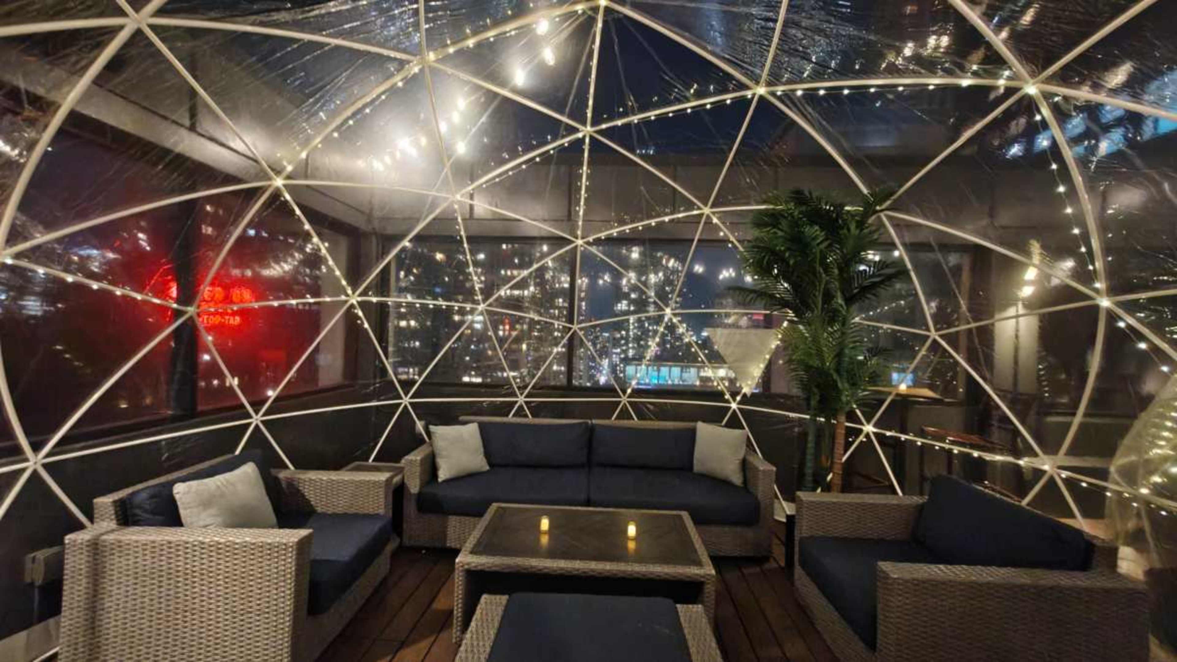 A clear igloo-style structure with seating and decorative lights offers a view of a city skyline at night.