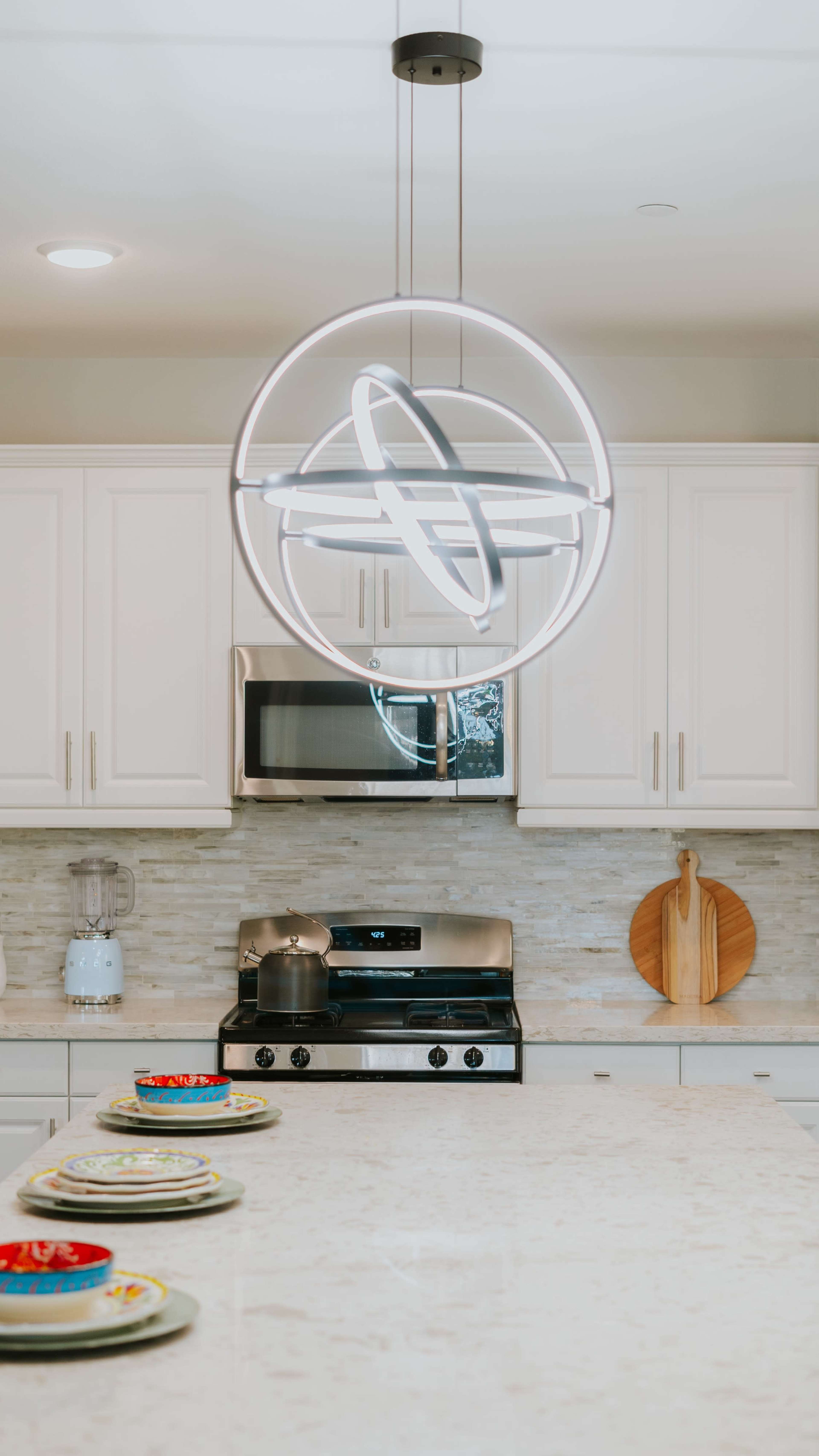 A modern kitchen features white cabinets, a stainless steel stove, a circular pendant light, and a marble countertop with dishes arranged neatly.
