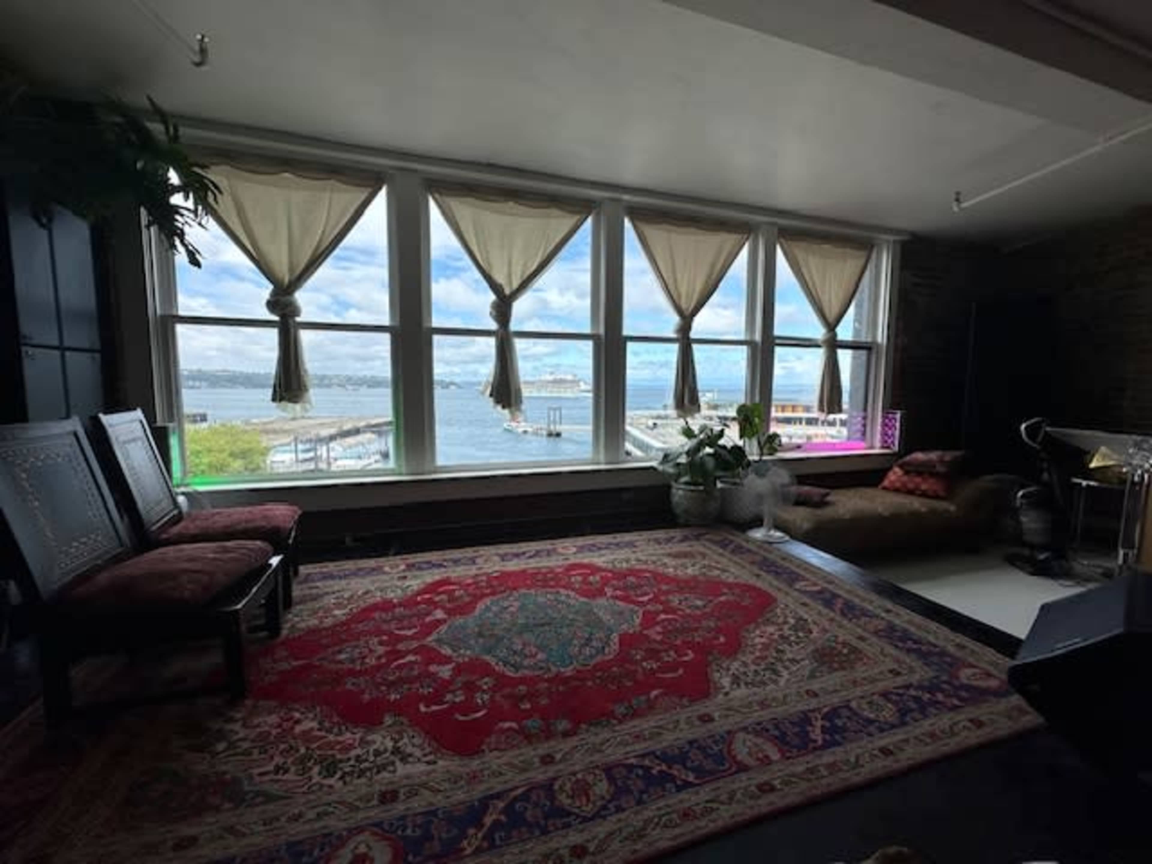 A cozy room features large windows with curtains, overlooking a waterfront view and decorated with a colorful rug and furniture.