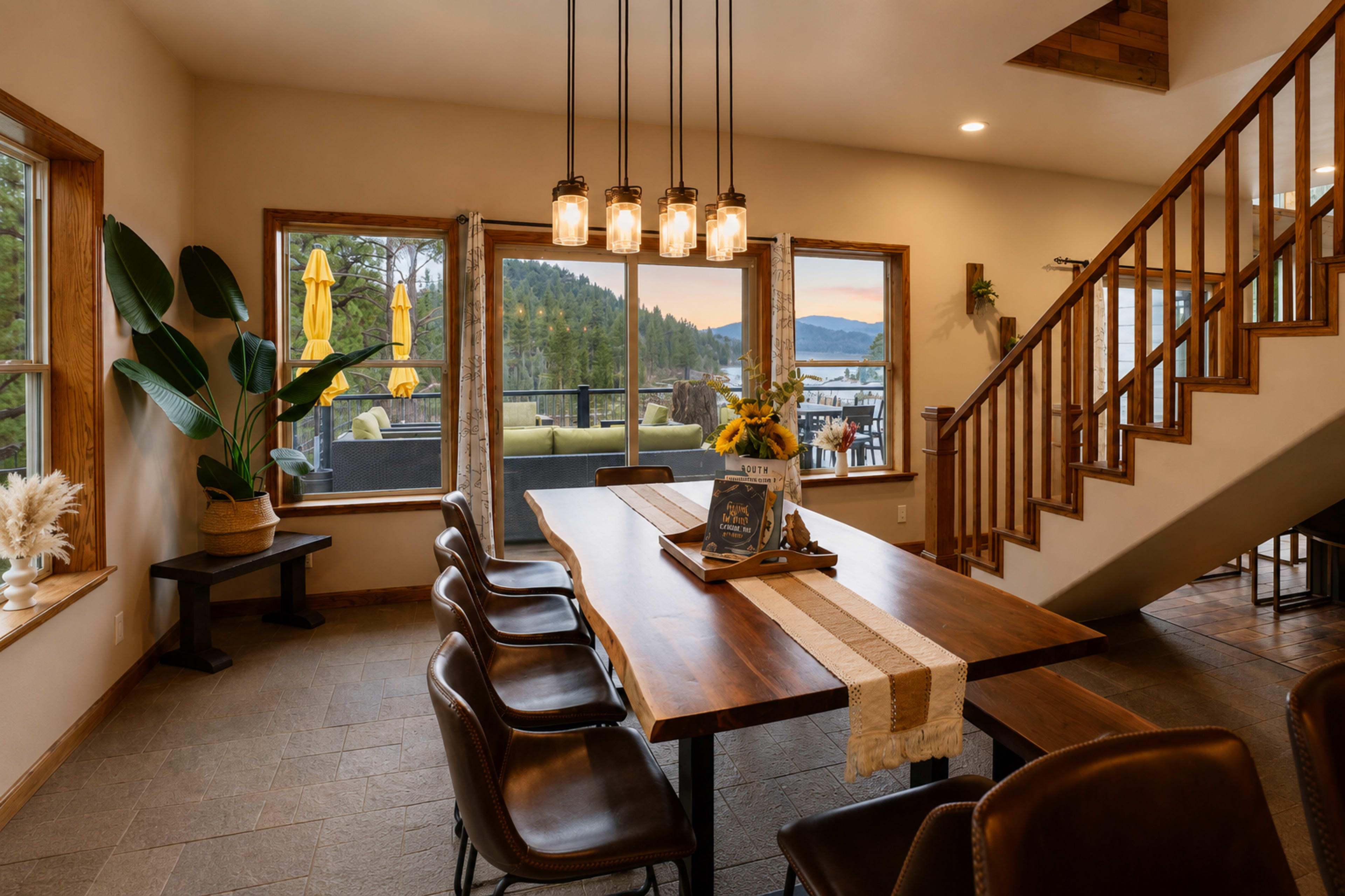 The image features a modern dining area with a large wooden table, surrounded by dark leather chairs, and large windows offering a view of a lake and mountains.