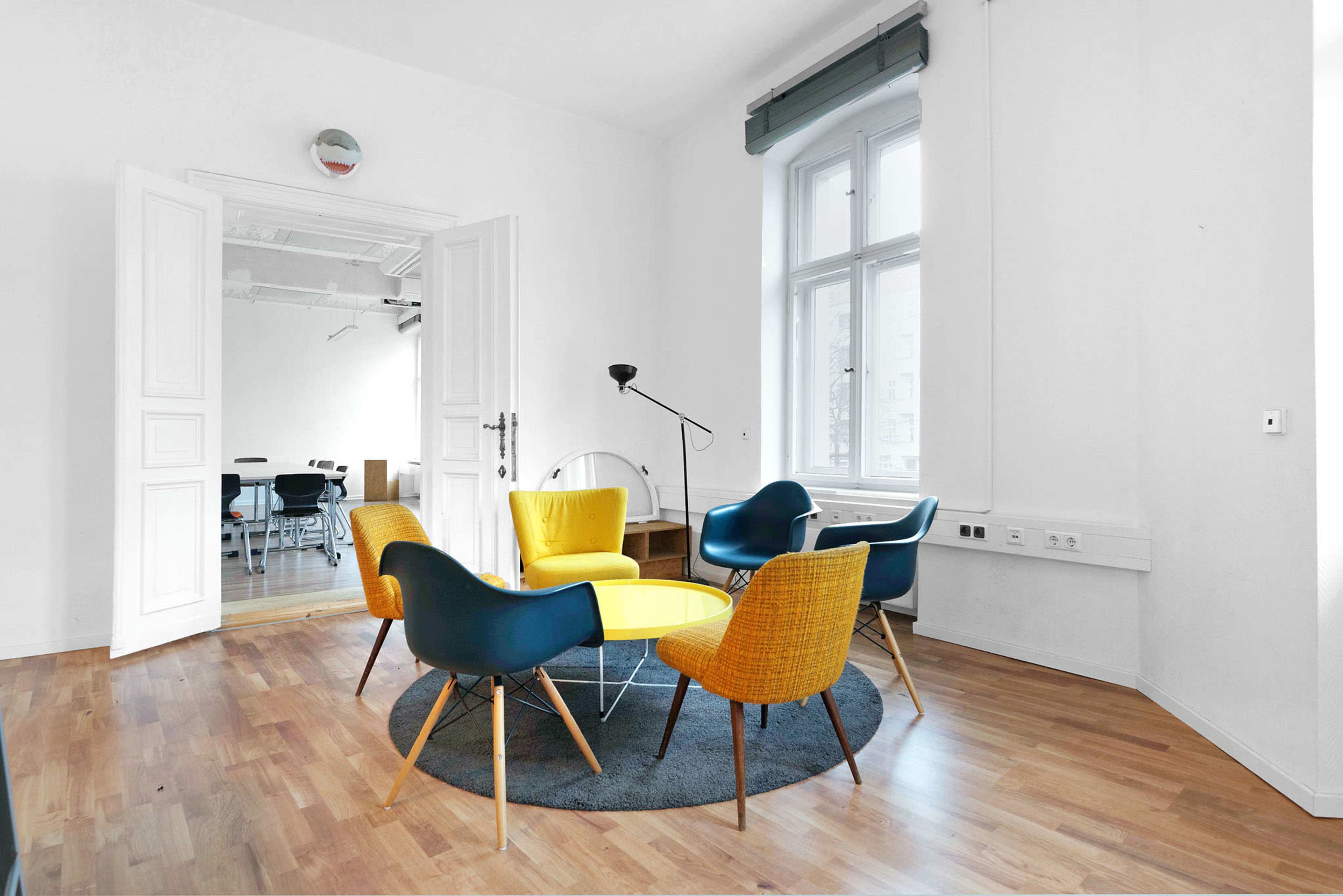 A bright, minimalist room features four chairs with colorful seats arranged around a round table on a dark rug.