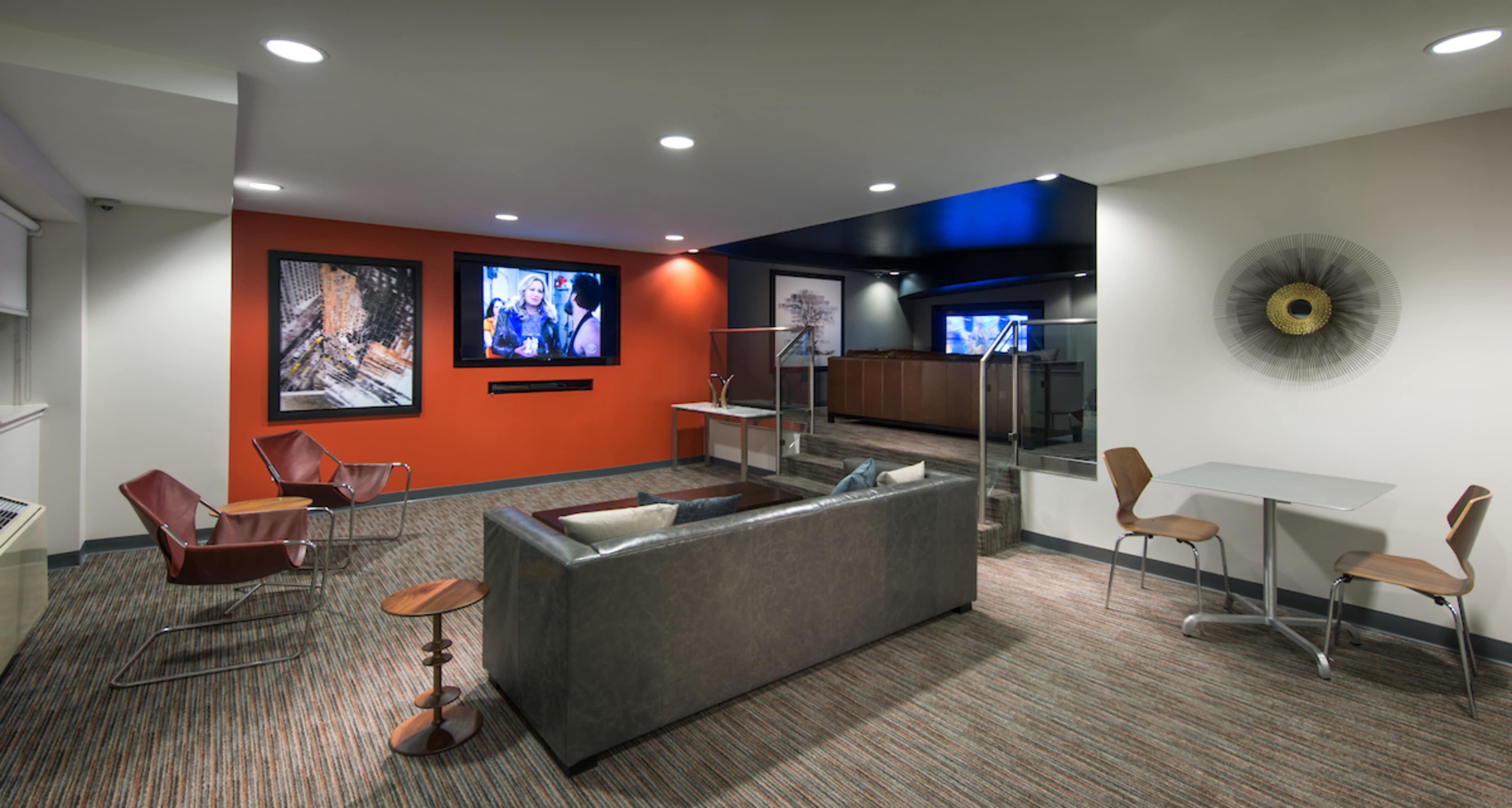 The image shows a modern lounge area with a sofa, armchairs, and a large screen displaying a video, set against a backdrop of contrasting colored walls and a reception area in the background.
