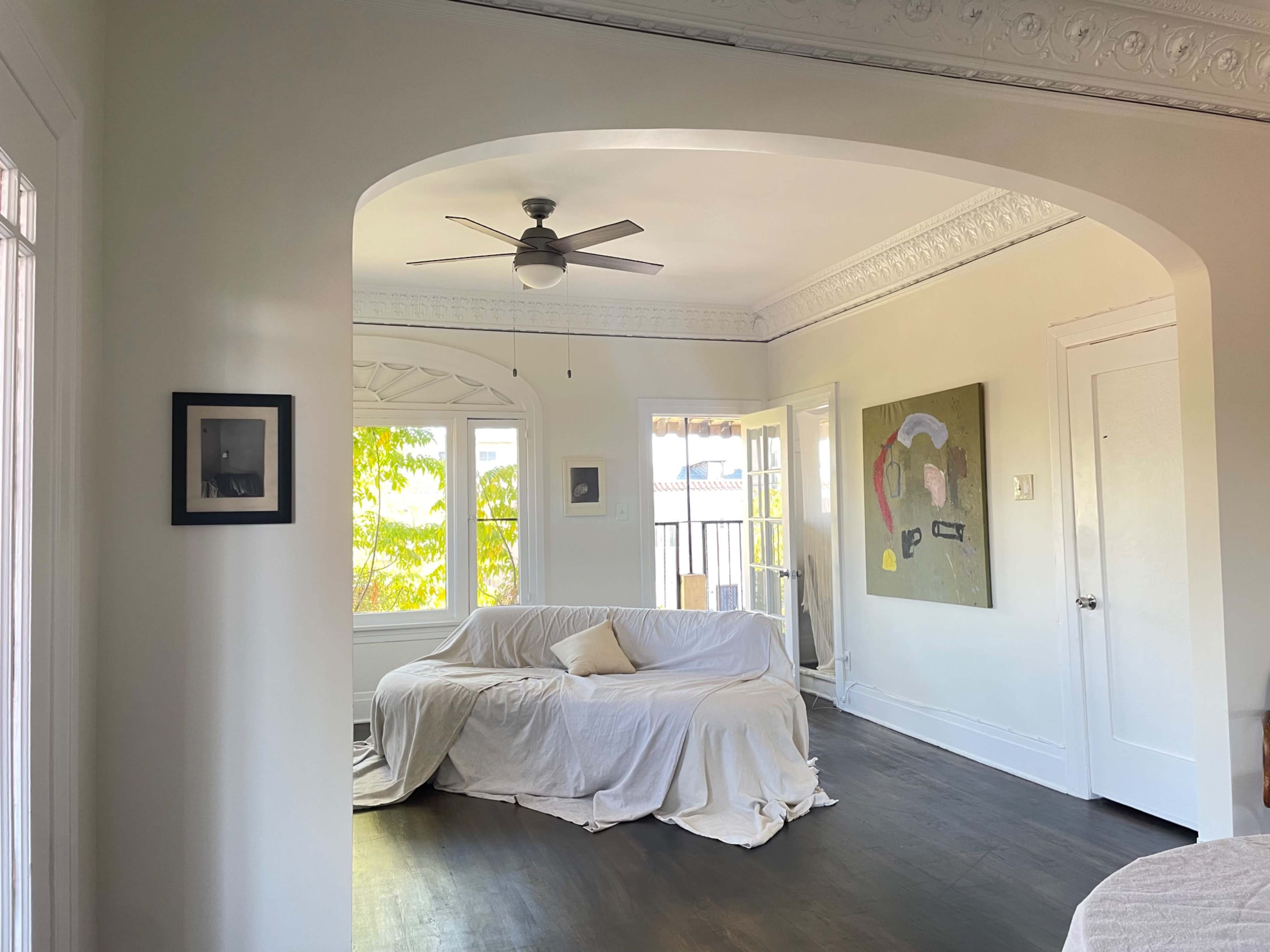 The image shows a bright living room featuring a white sofa draped with a cover, a ceiling fan, and large windows providing natural light.