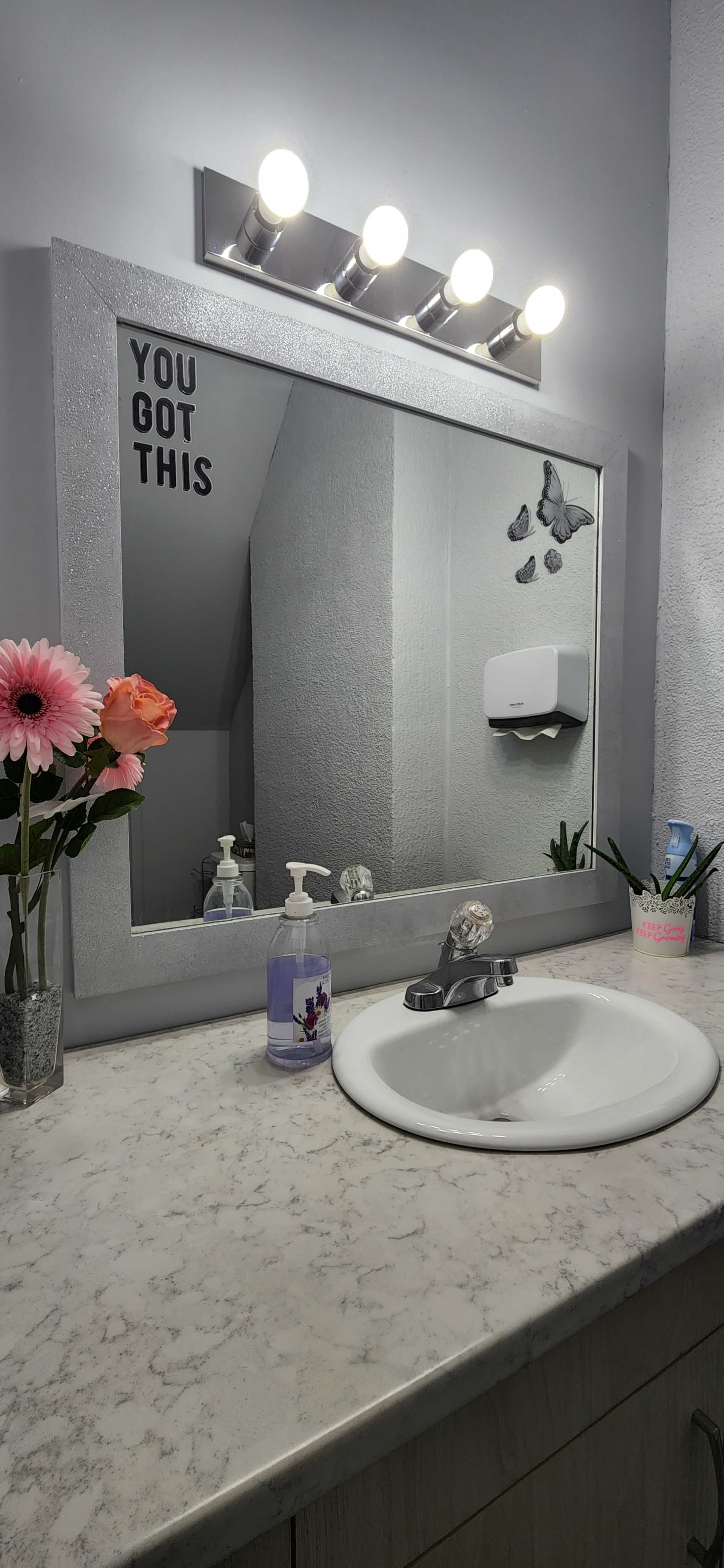 A bathroom vanity with a mirror, a floral arrangement, soap dispenser, and a wall-mounted paper towel holder, featuring the phrase "YOU GOT THIS" on the mirror.