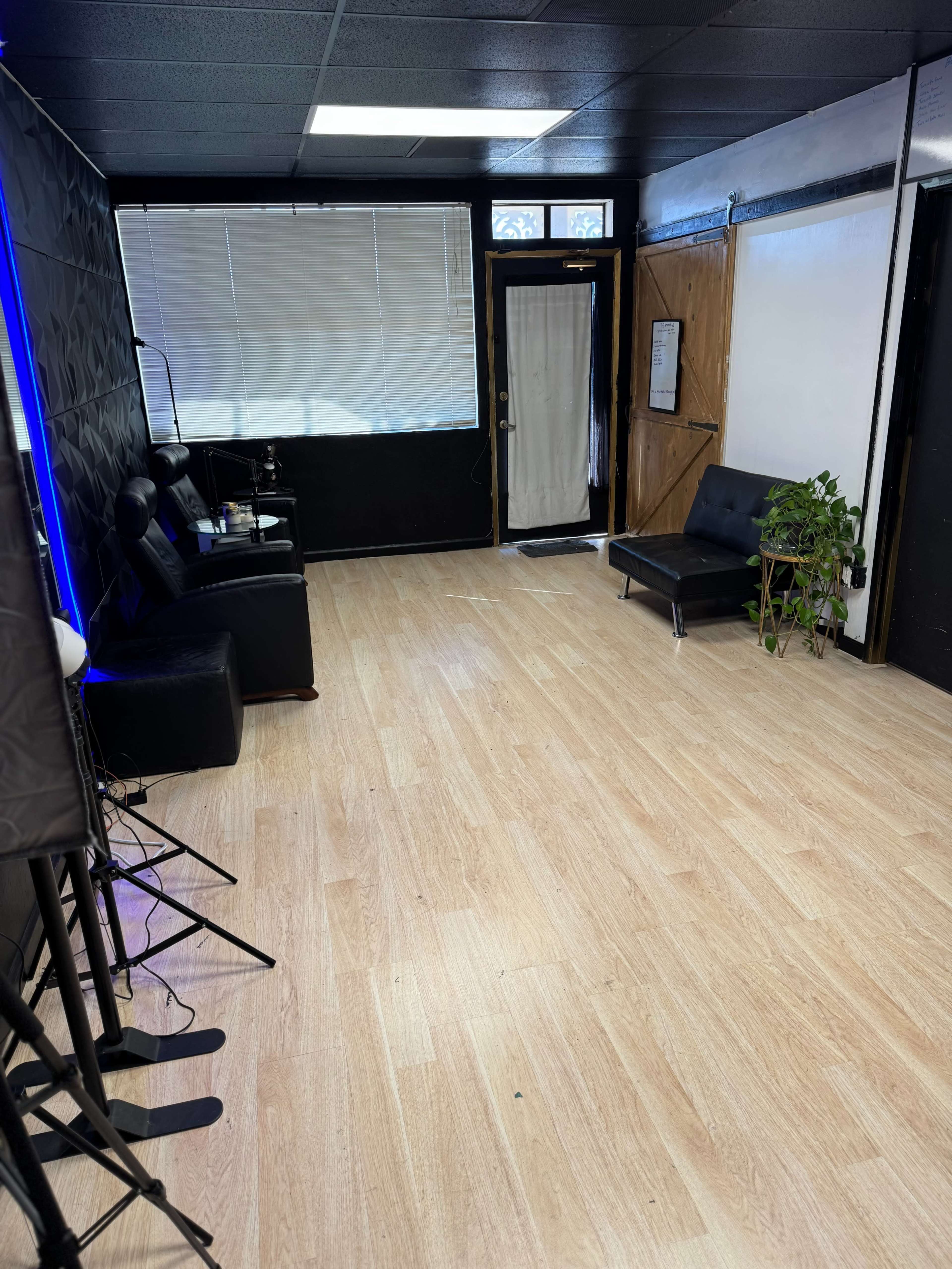 The image shows a sparsely furnished interior space with light wooden flooring, a door leading outside, black walls, and a few seating options.