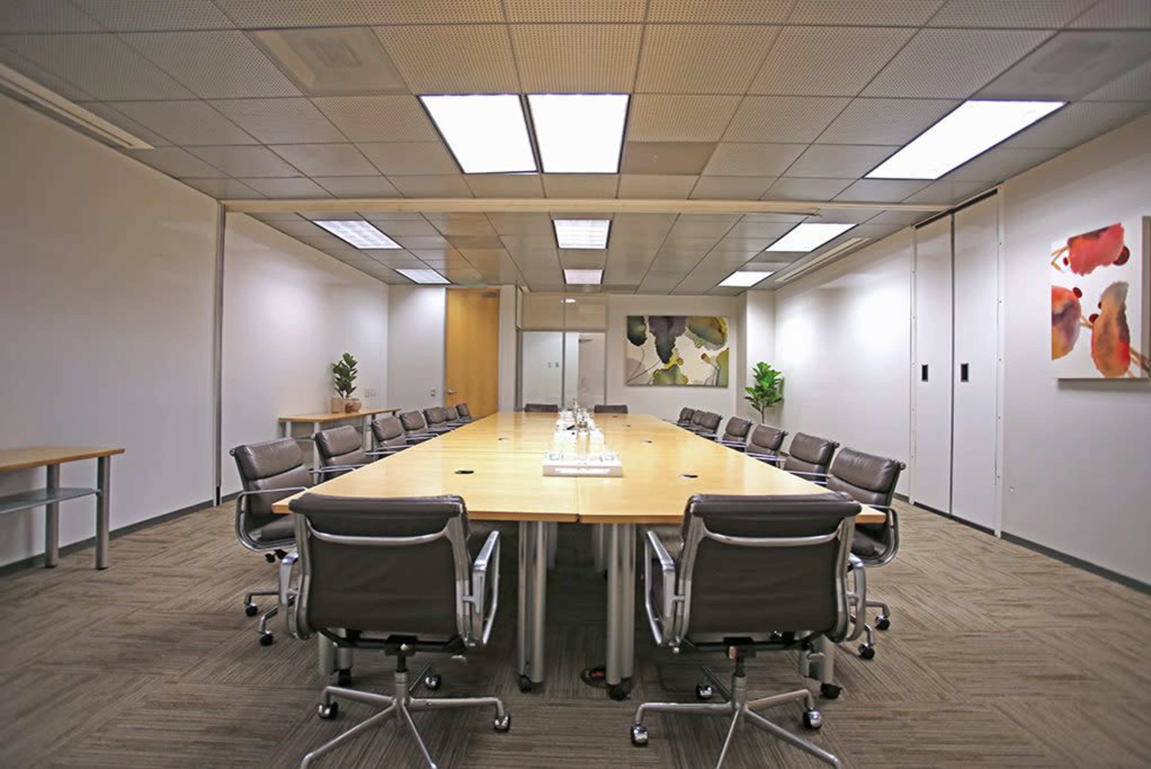 A large, empty conference room features a long wooden table, grey office chairs, and abstract artwork on the walls.