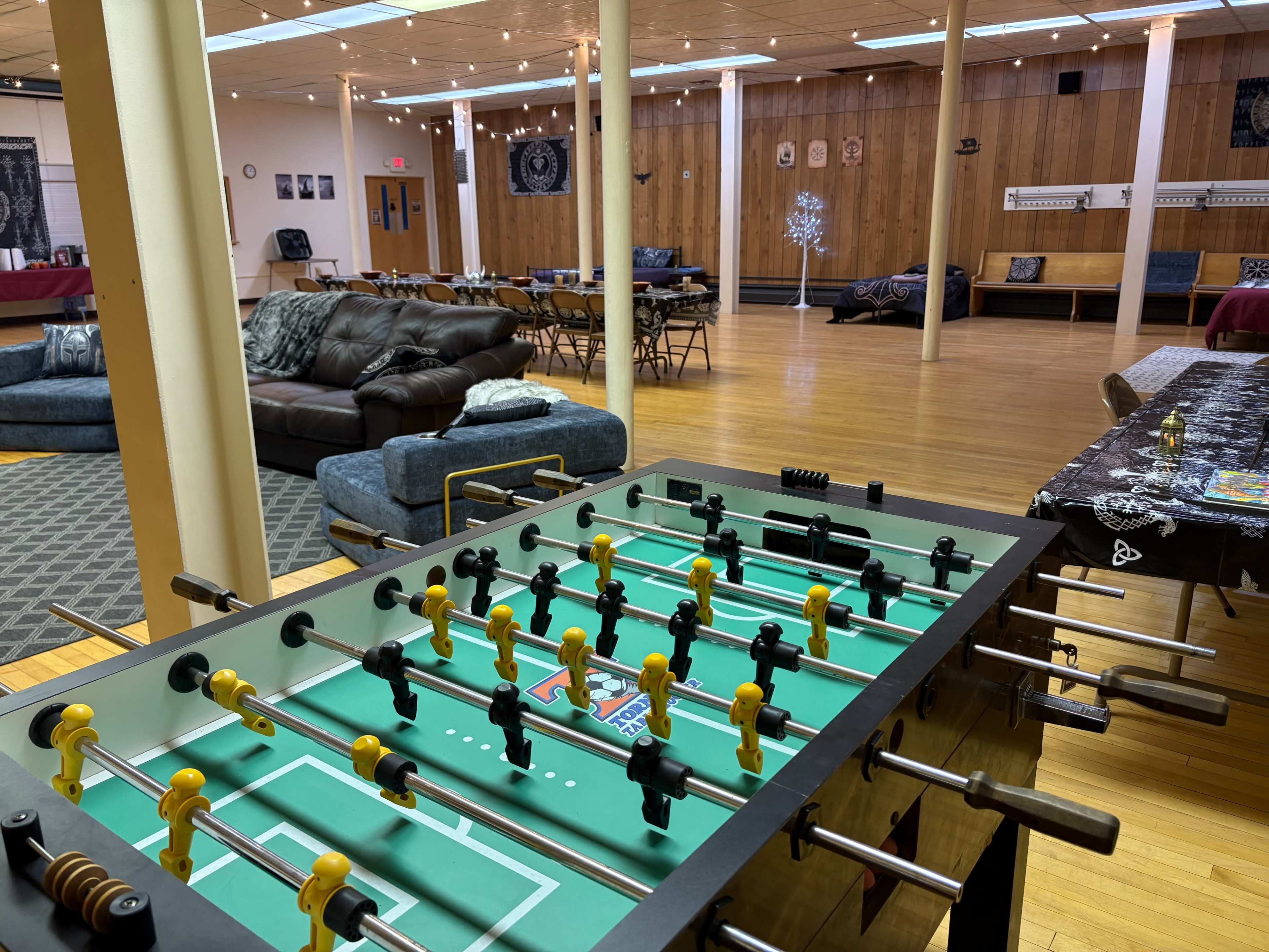 The image shows a spacious common room with a foosball table in the foreground, couches, tables, and a wooden wall in the background.