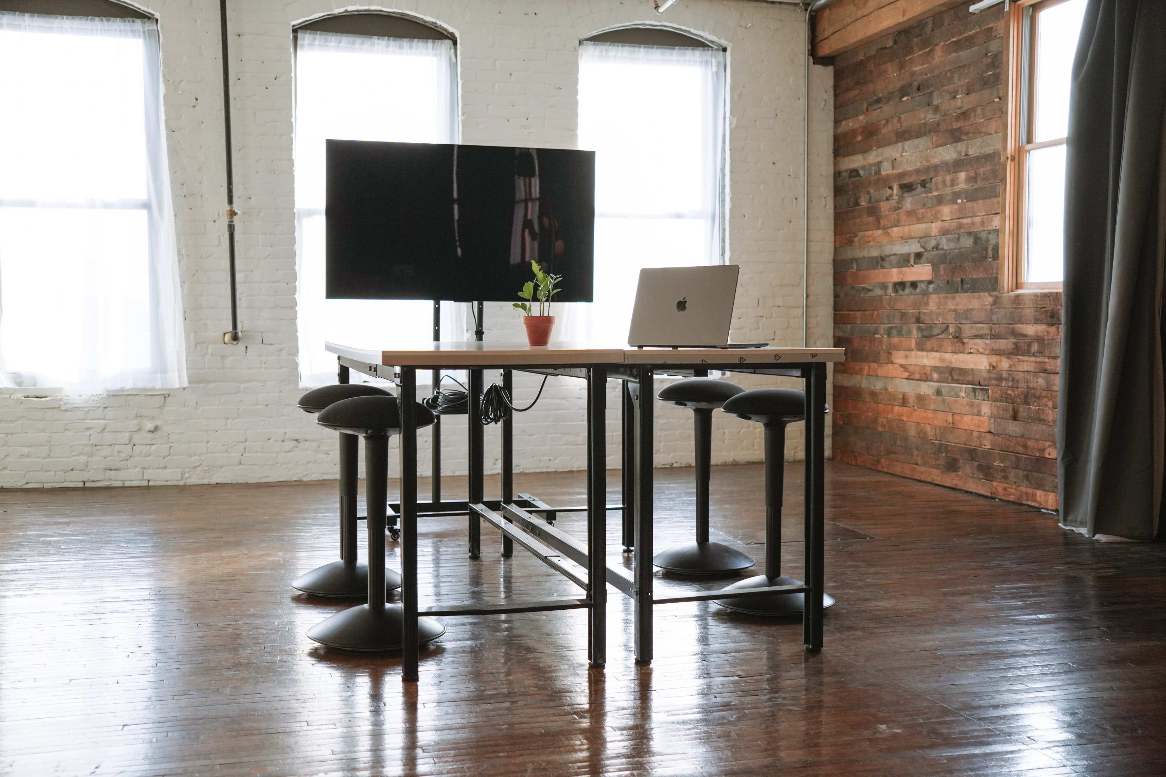 A work desk with a laptop and a potted plant stands beside a large monitor in a spacious, well-lit room with wooden walls.