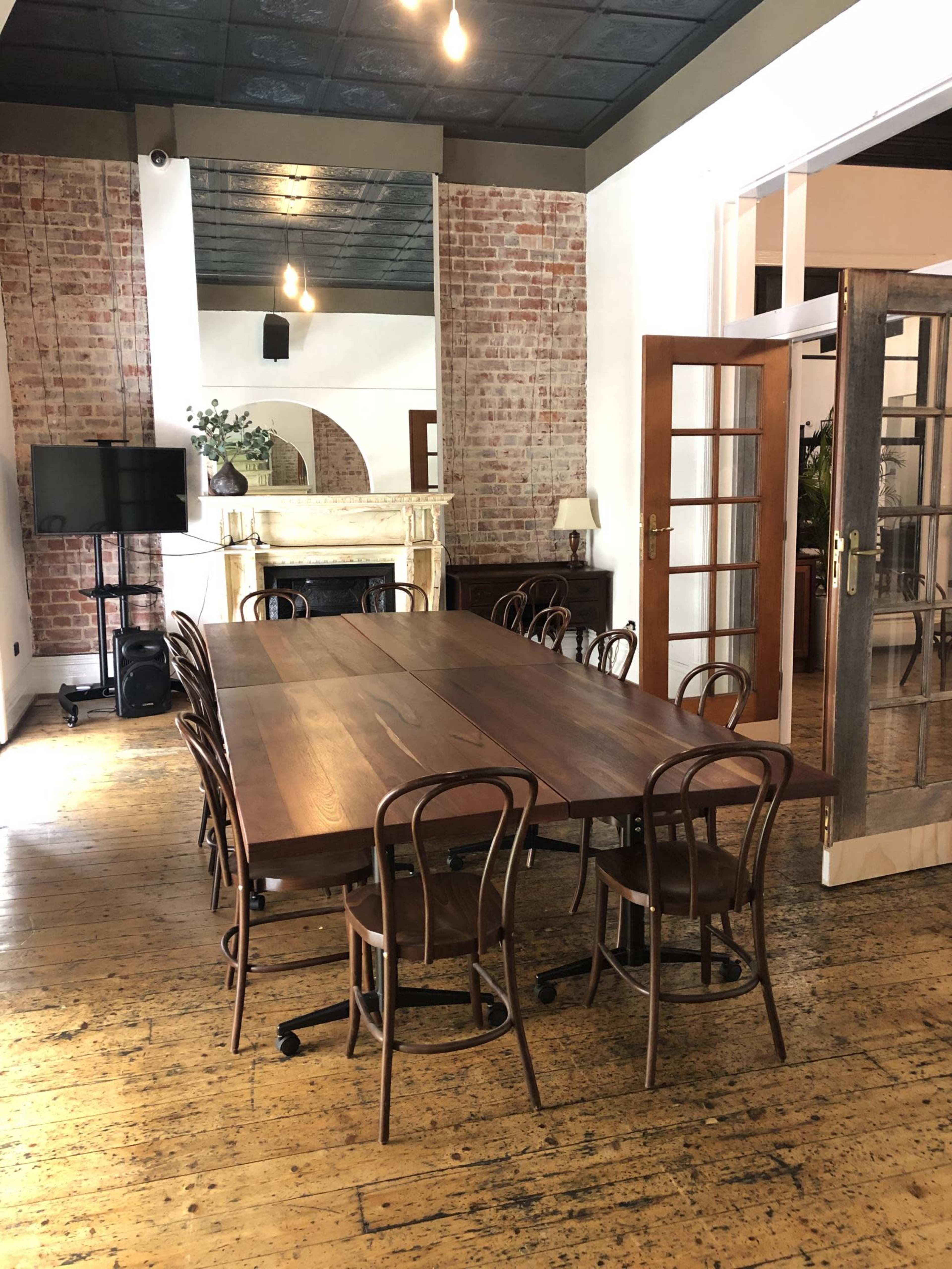 A large wooden table with rolling chairs is set in a room featuring exposed brick walls and a vintage fireplace.
