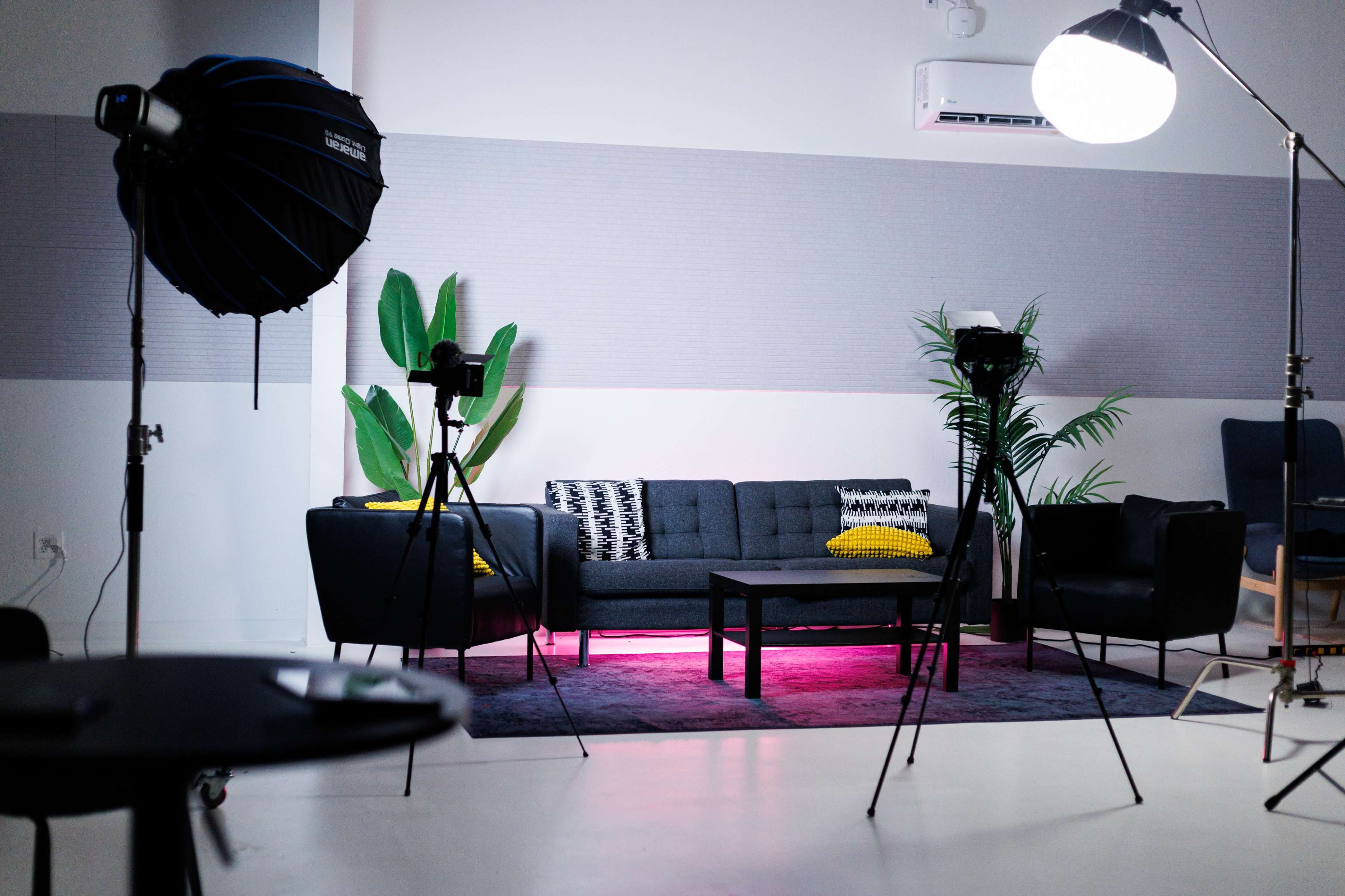 The image features a modern photo studio setup with a gray sofa, accent chairs, and lighting equipment arranged around a low table on a patterned rug.