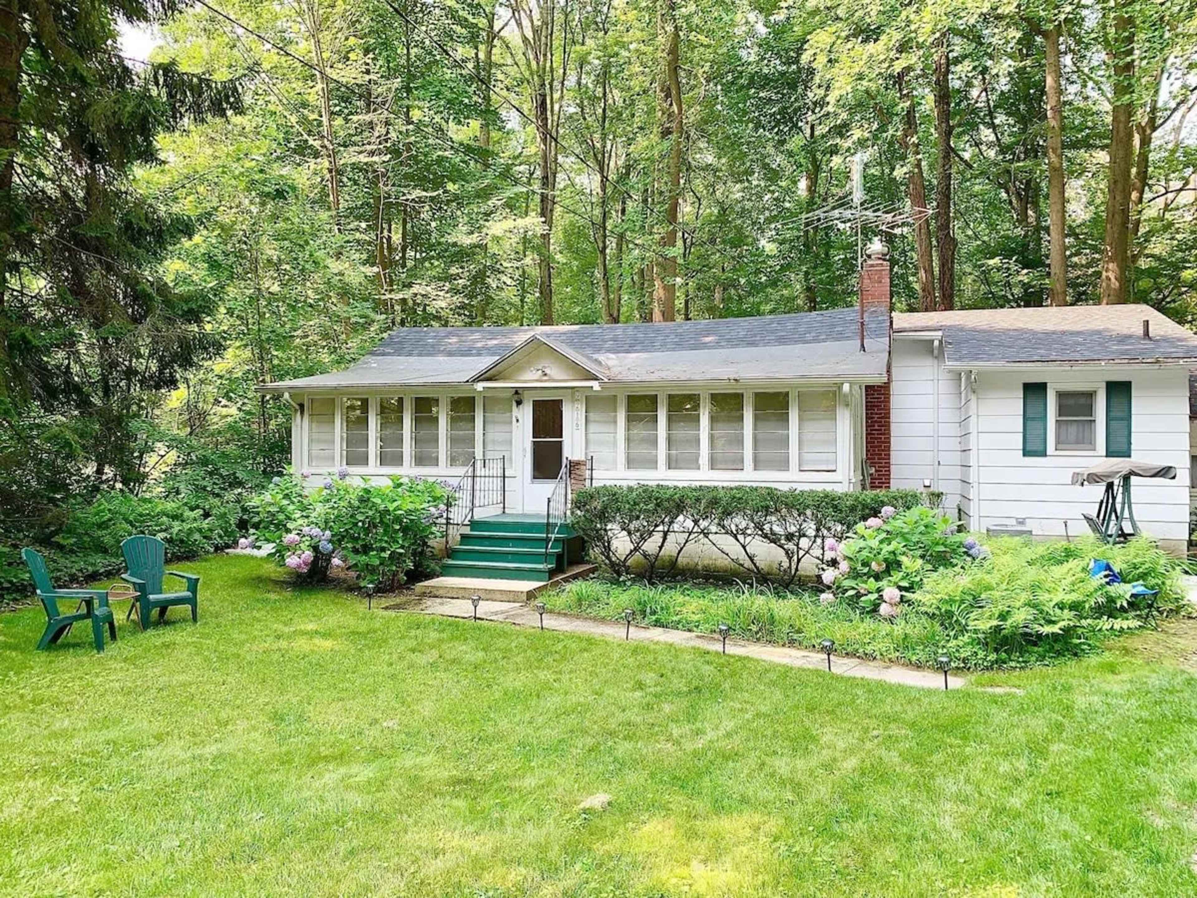 A single-story house with a front porch is set in a wooded area, surrounded by greenery and blooming plants.