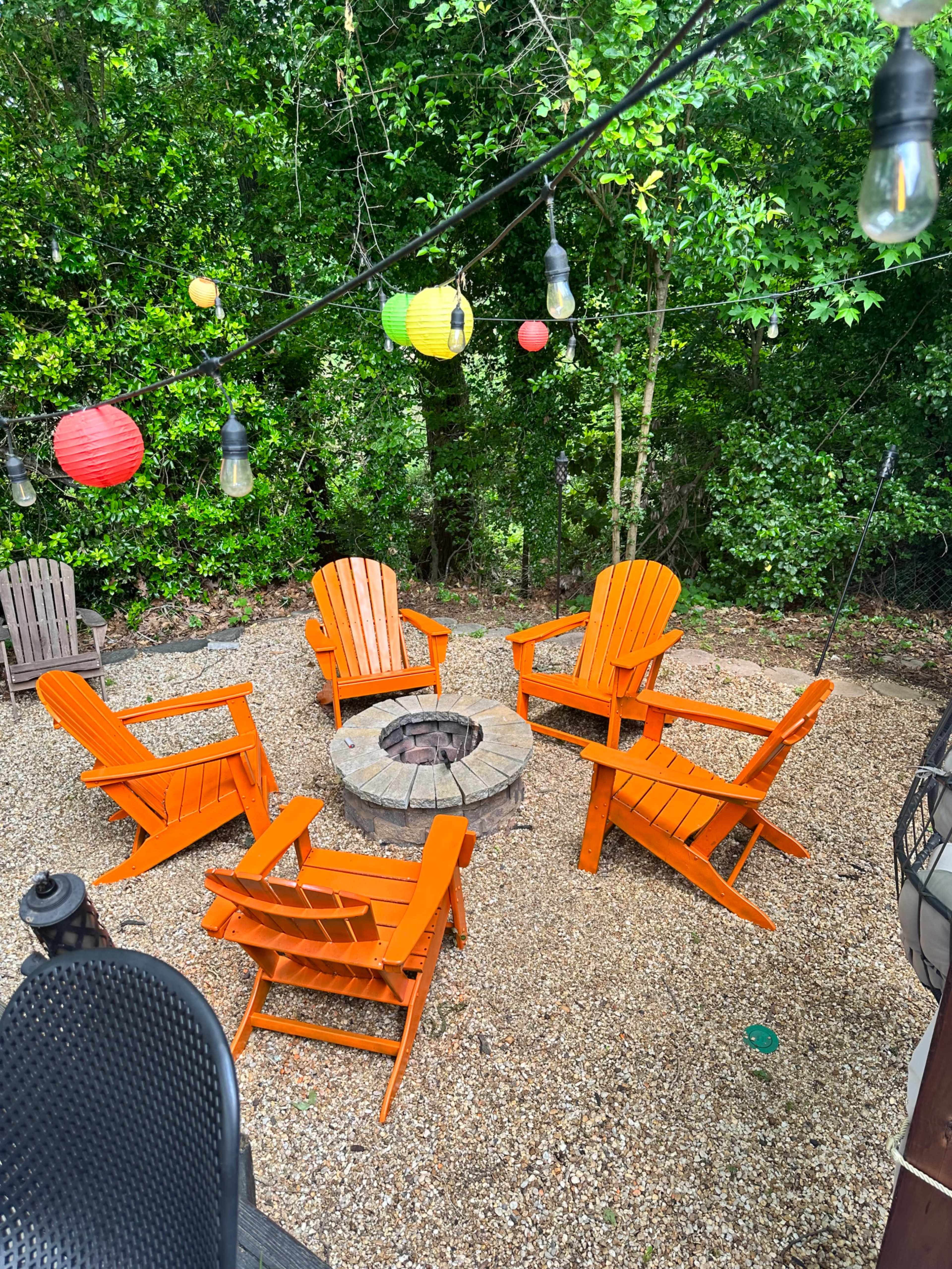 A circular arrangement of five orange Adirondack chairs surrounds a stone fire pit in a gravel outdoor area, accented by hanging lanterns and surrounded by greenery.