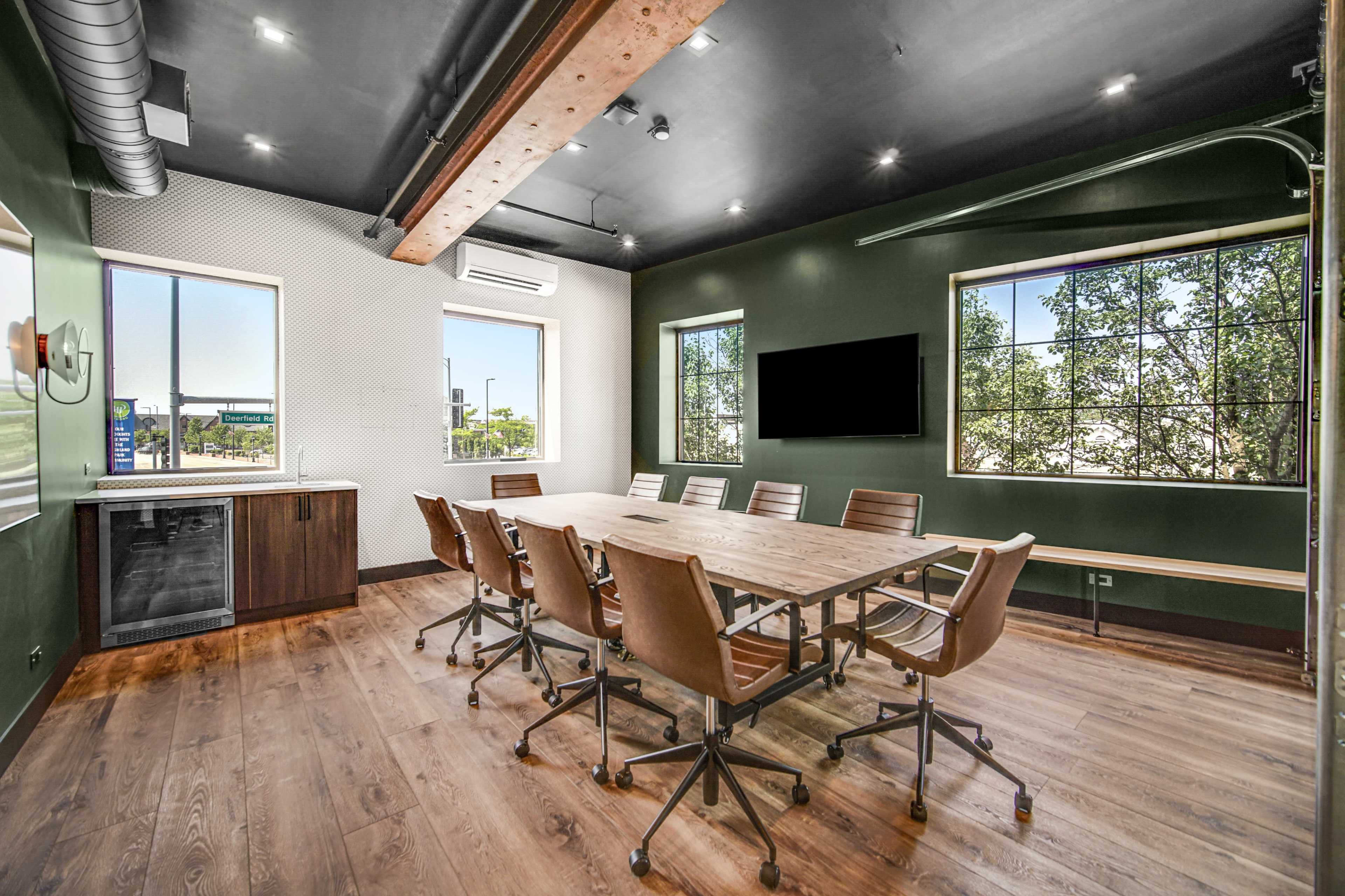 A conference room features a large wooden table surrounded by leather chairs, with windows providing natural light and a wall-mounted television.