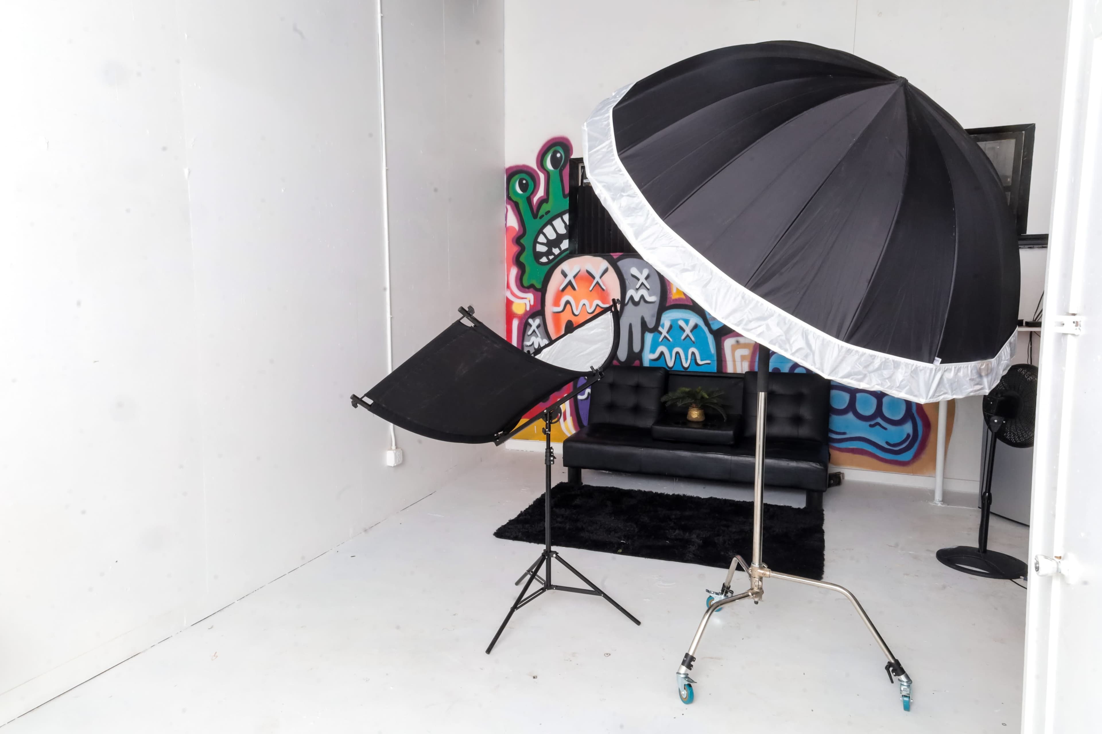 The image shows a photo studio setup featuring a large black umbrella light and a reflector stand in front of a black couch against a colorful graffiti wall.