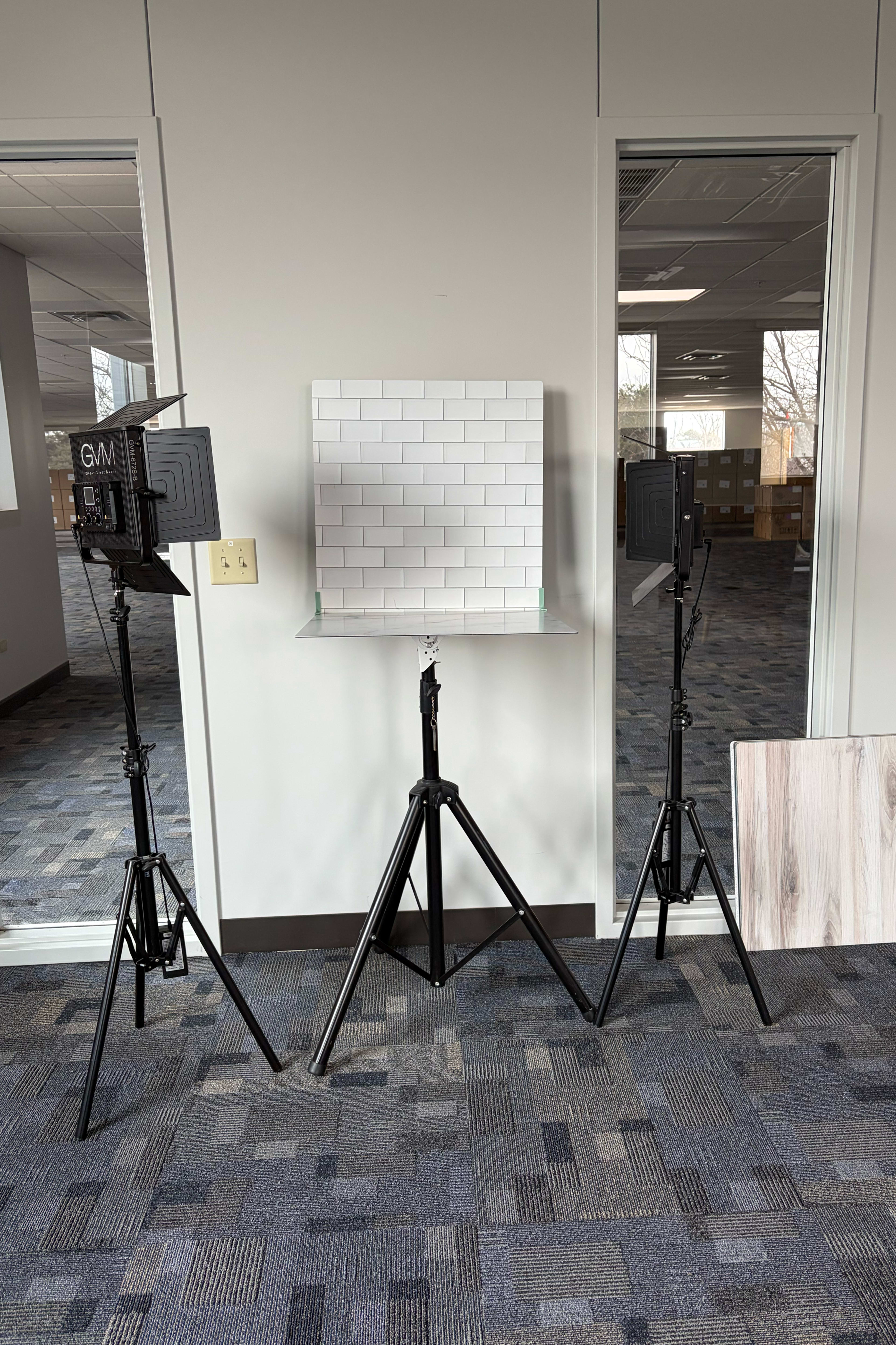 The image displays a setup for photography, featuring two adjustable lights and a square tile backdrop on a transparent platform, all situated in a room with gray carpet and large windows.