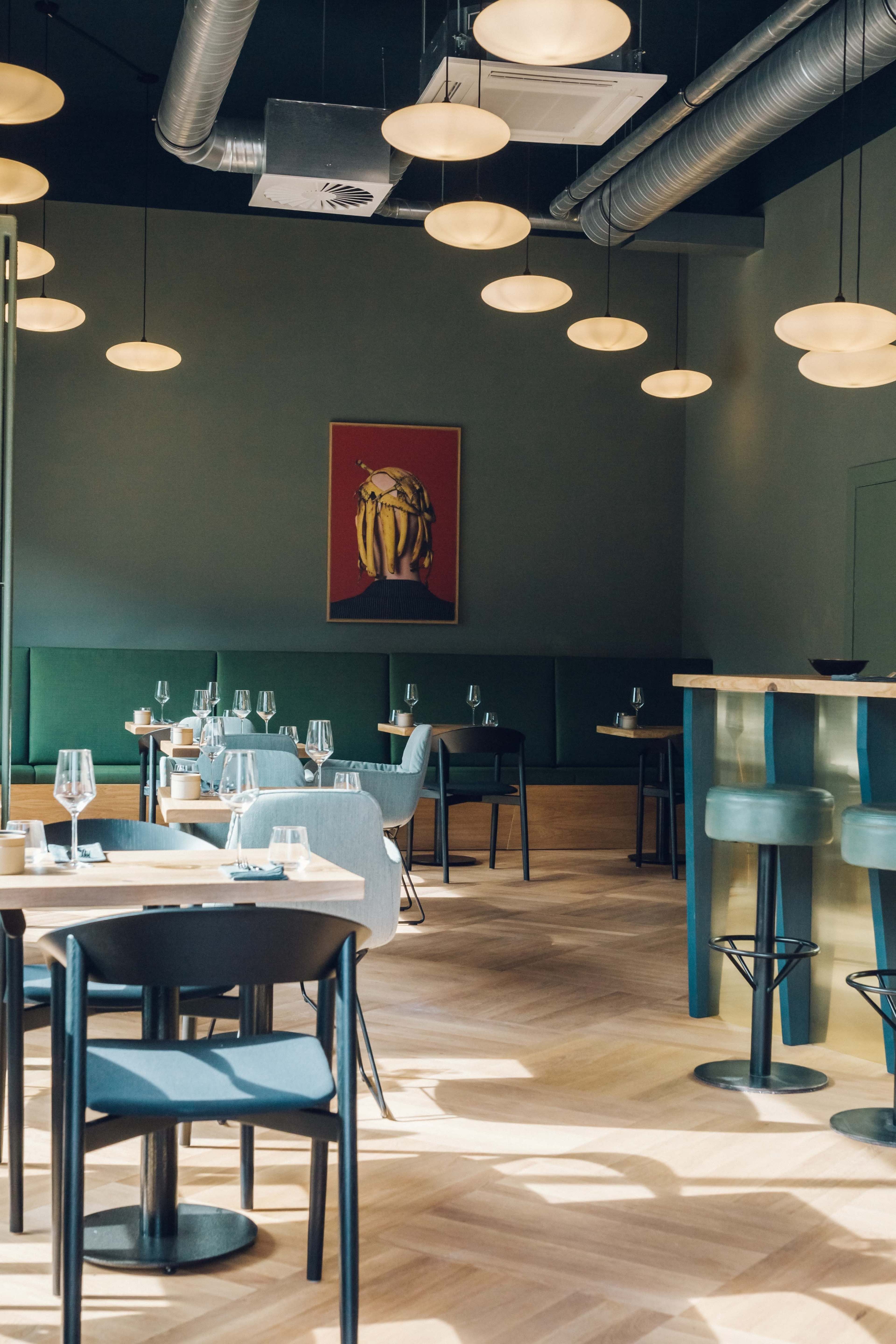 The image shows an upscale restaurant interior with modern furniture, wall art, and an open layout featuring tables and a bar area.