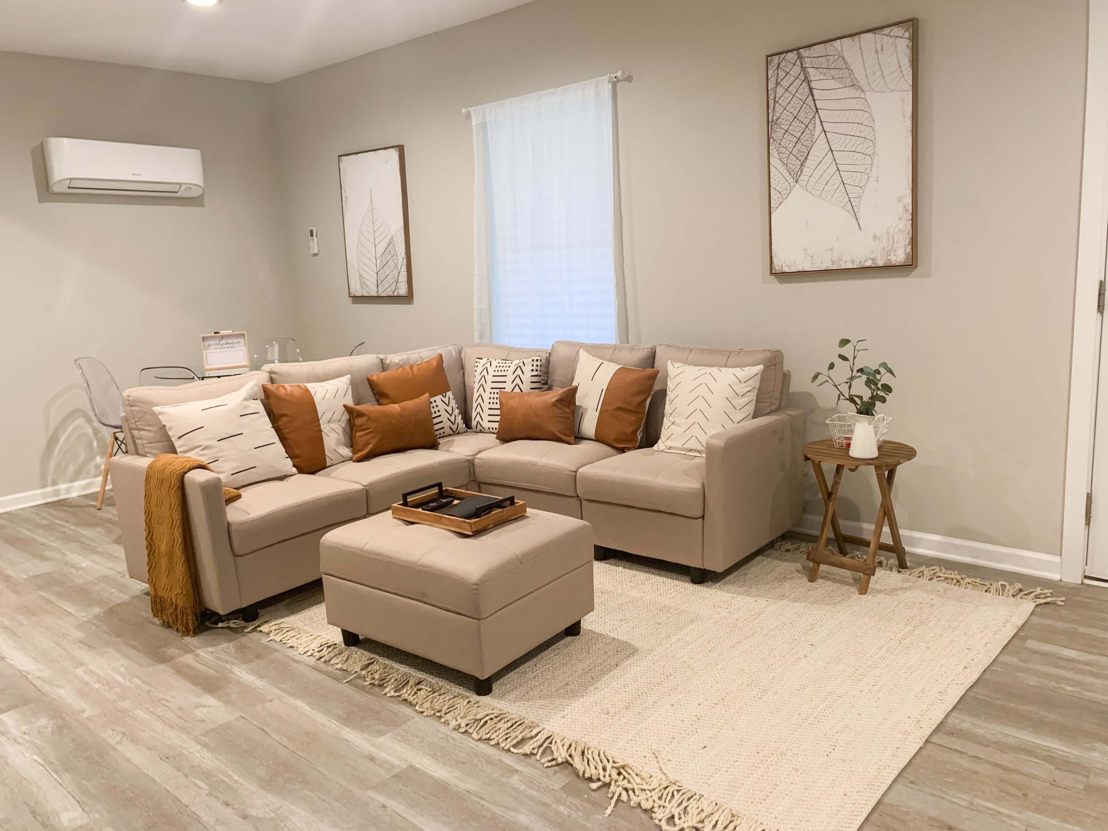 A light-colored living room featuring a sectional sofa with patterned cushions, a small ottoman, a woven area rug, and two framed leaf-themed artworks on the walls.
