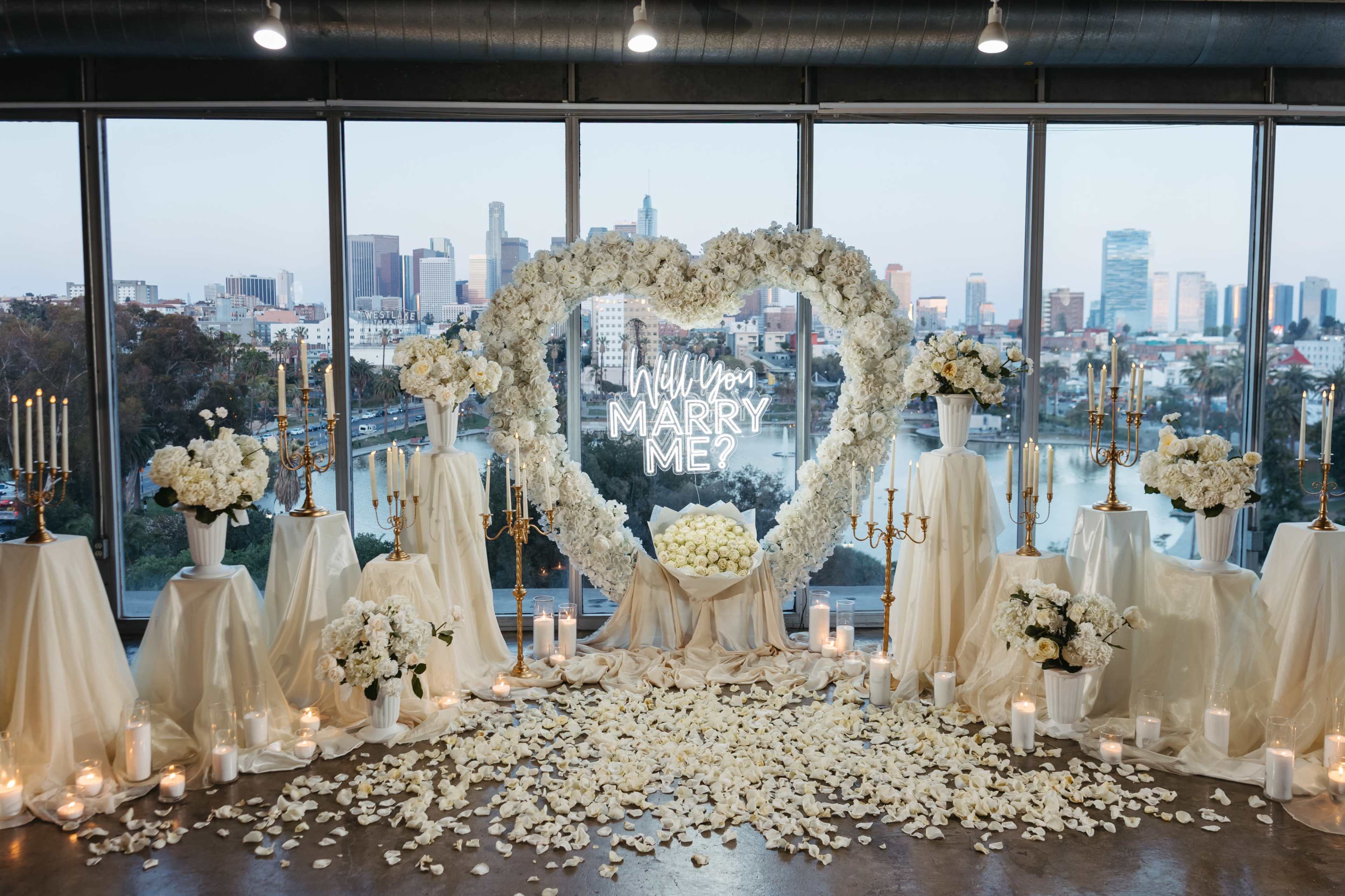 The image shows a romantic proposal setup featuring a heart-shaped flower arrangement with a glowing "Will You Marry Me?" sign, surrounded by white flowers and candles, all set against a city skyline.
