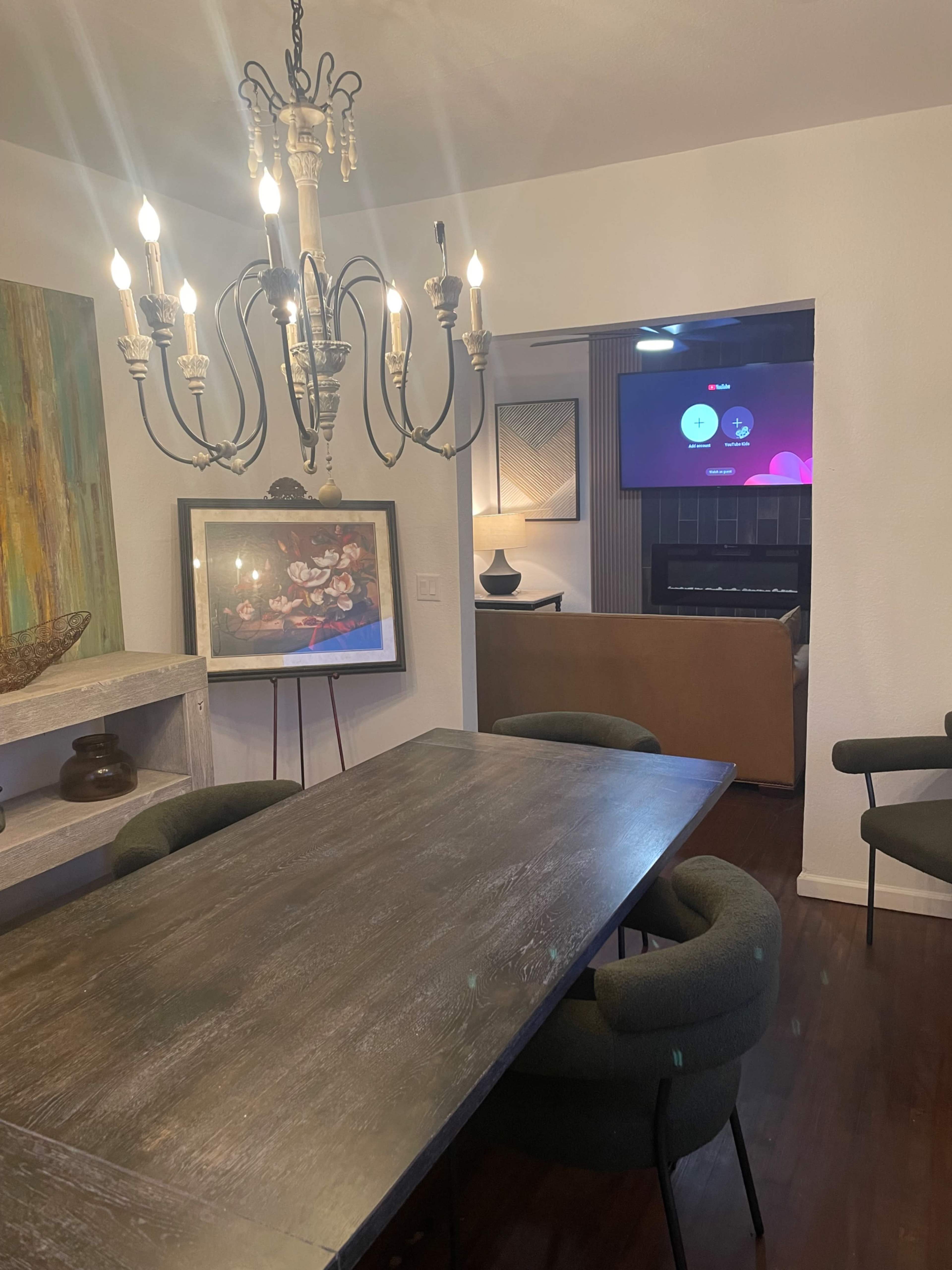 The image shows a dining room with a large wooden table, upholstered chairs, a chandelier above, and a living area in the background featuring a TV and a couch.