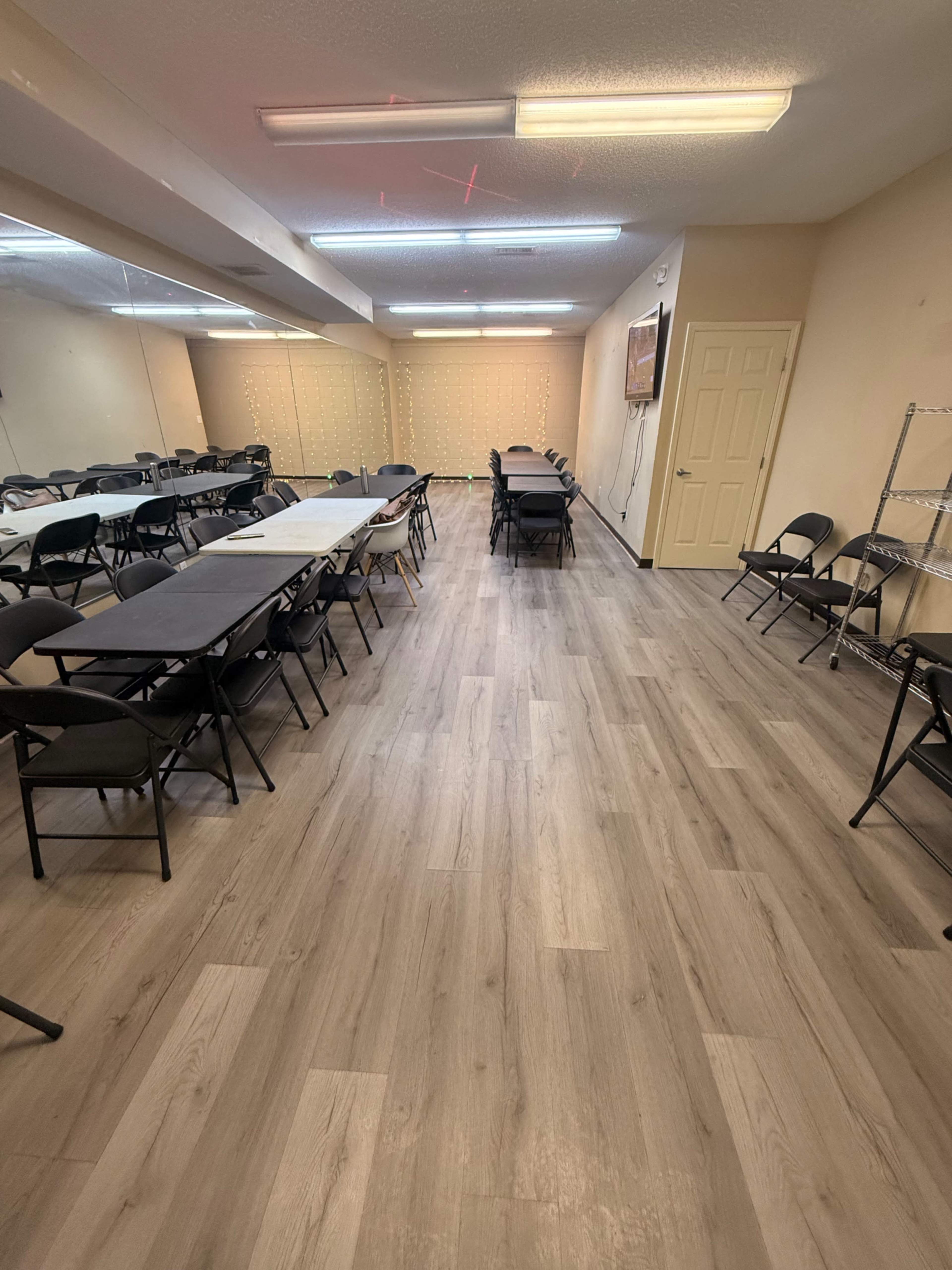 The image shows a spacious room with several rows of tables and chairs set up, along with a television mounted on the wall and a mirrored section on one side.