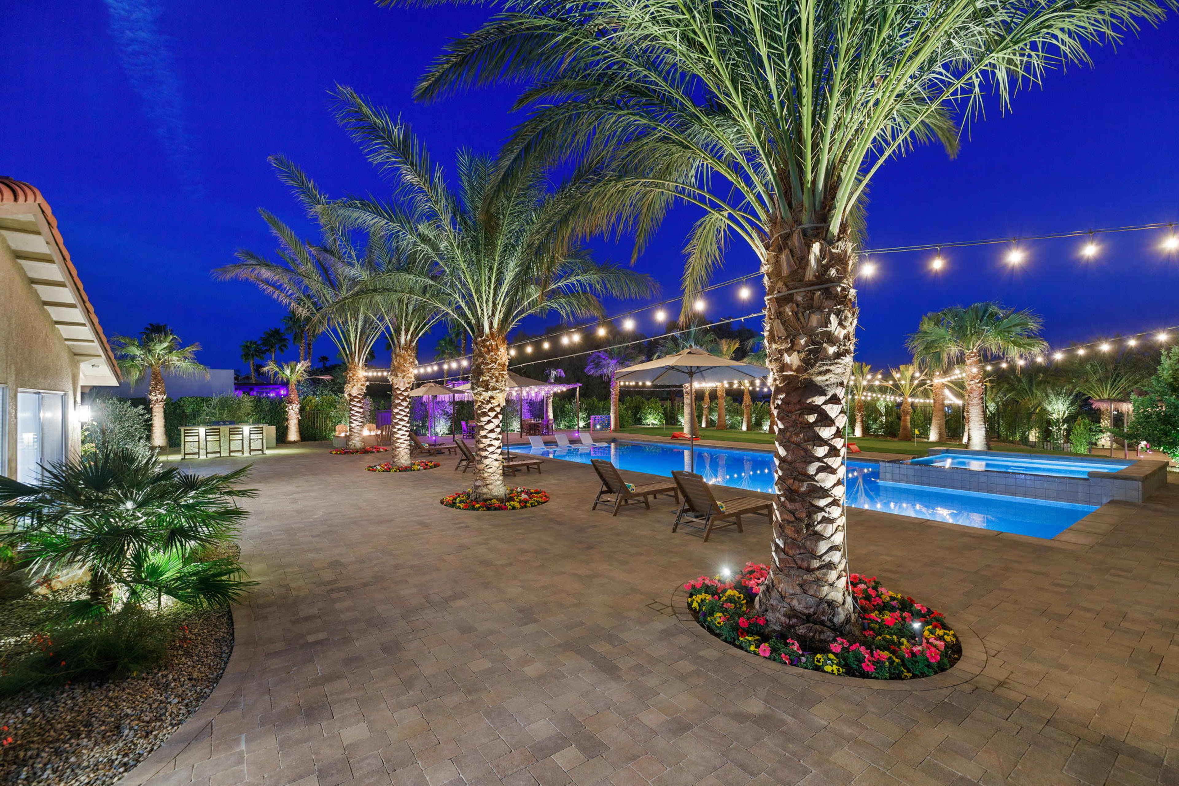 A landscaped pool area with palm trees, a swimming pool, and string lights under a nighttime sky.