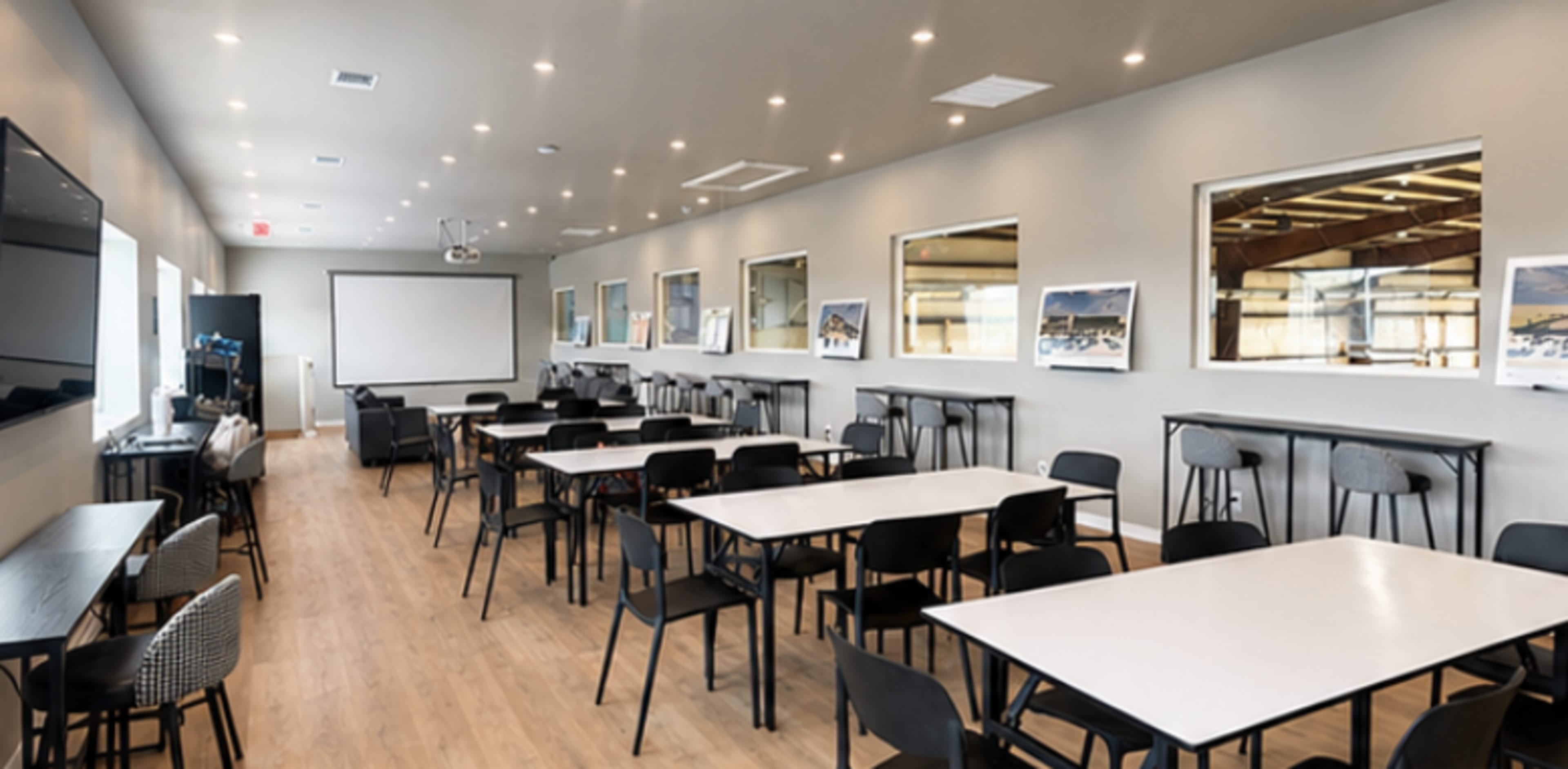 The image shows a spacious, brightly lit meeting room with tables and chairs arranged, featuring large windows and a projector screen at one end.