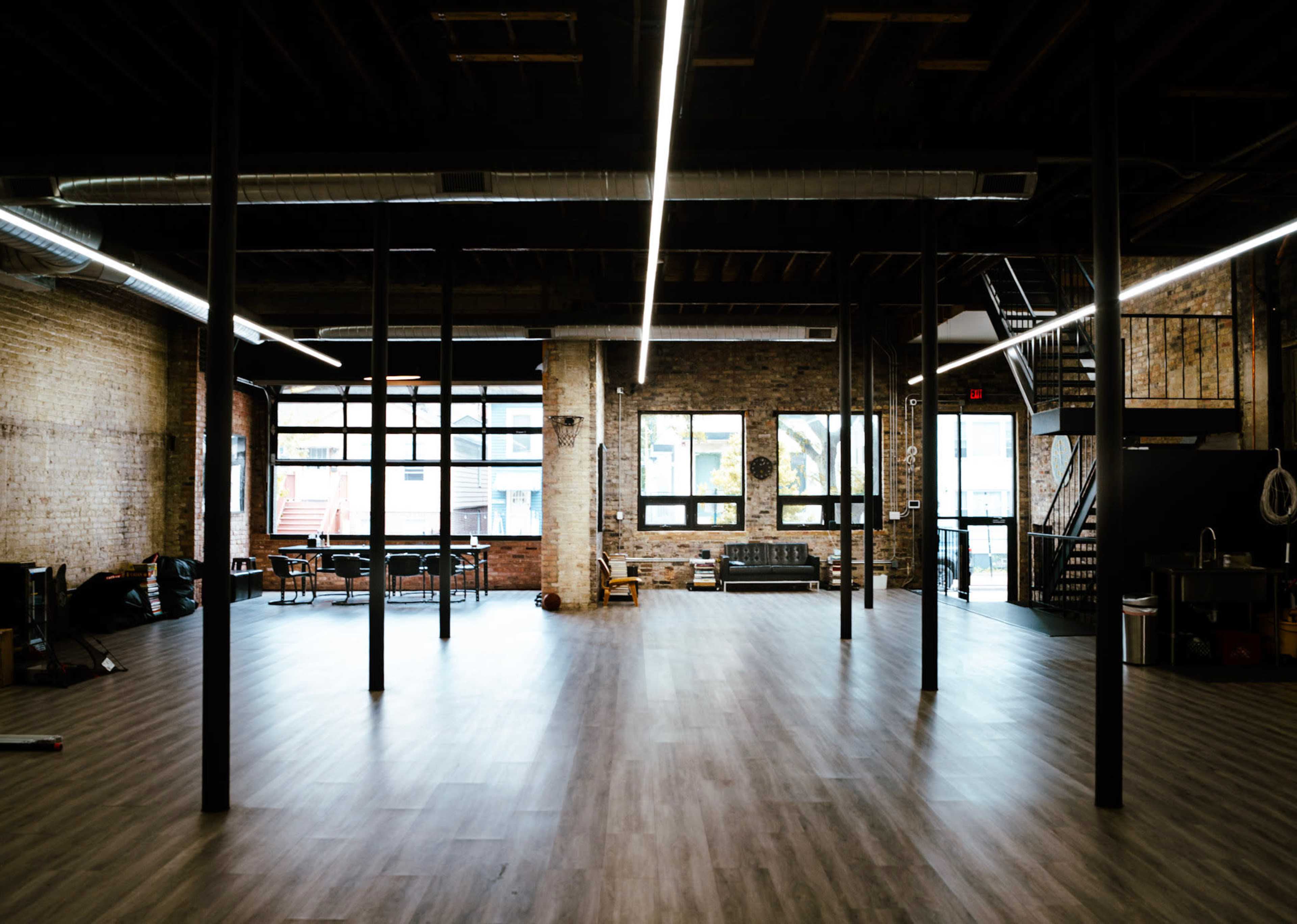 The image shows a spacious, industrial-style interior with exposed brick walls, wooden floors, and large windows, featuring several support columns and seating areas.