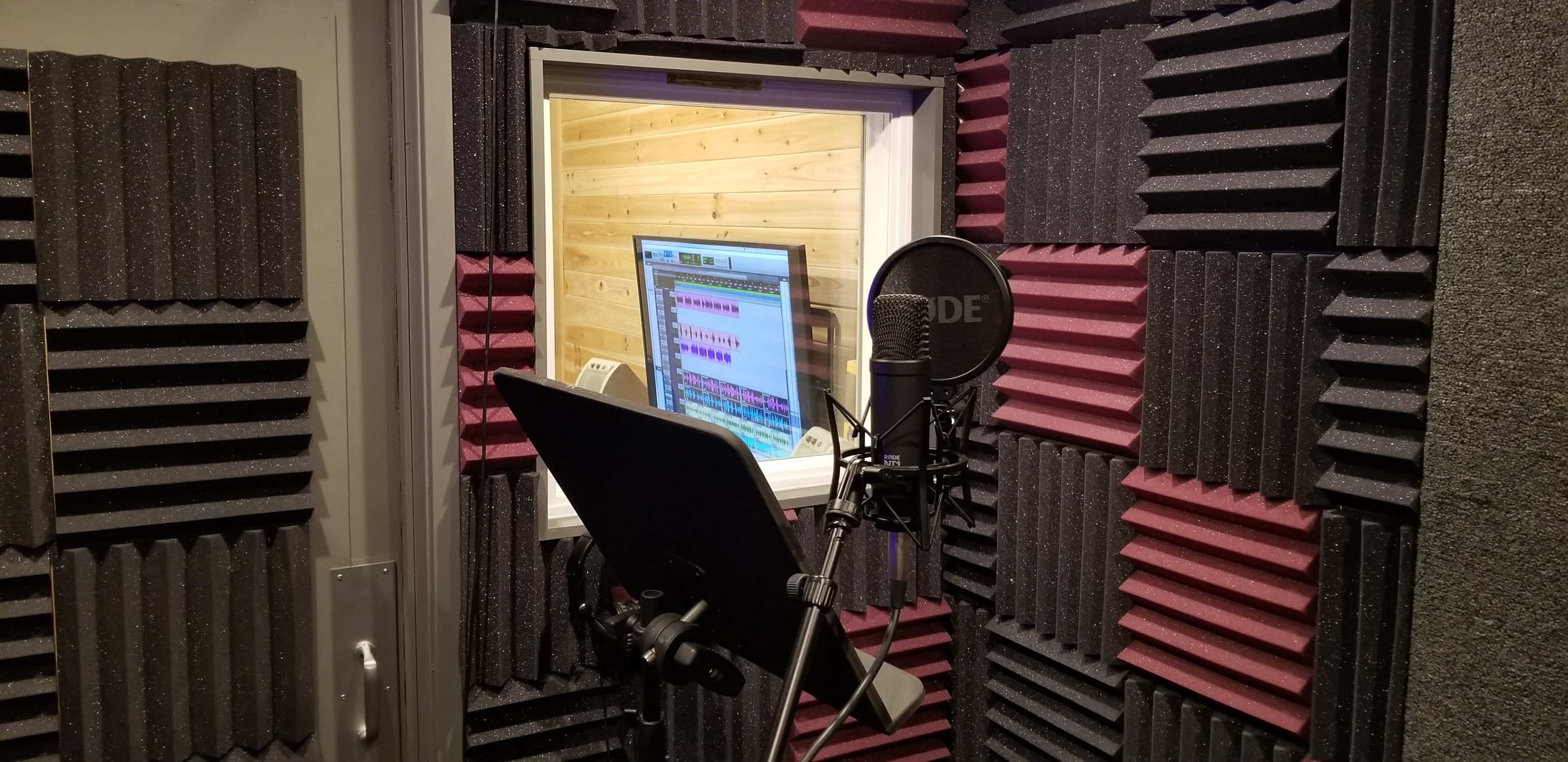 A sound booth with acoustic foam panels on the walls, a microphone on a stand, and a monitor visible through a window.