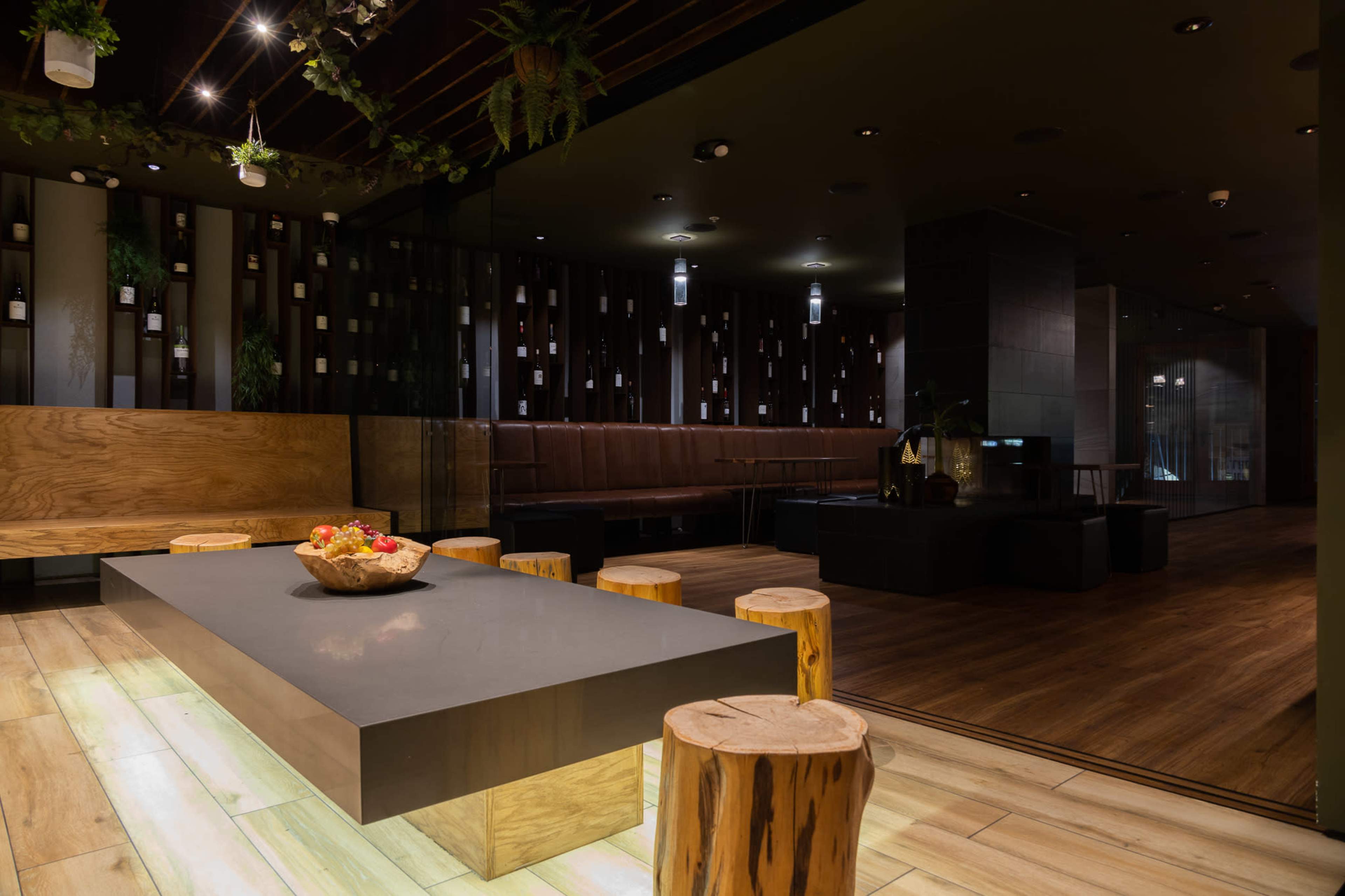 The image shows a modern interior space featuring a large gray table surrounded by wooden stools, with an ambiance created by dark walls and ambient lighting.