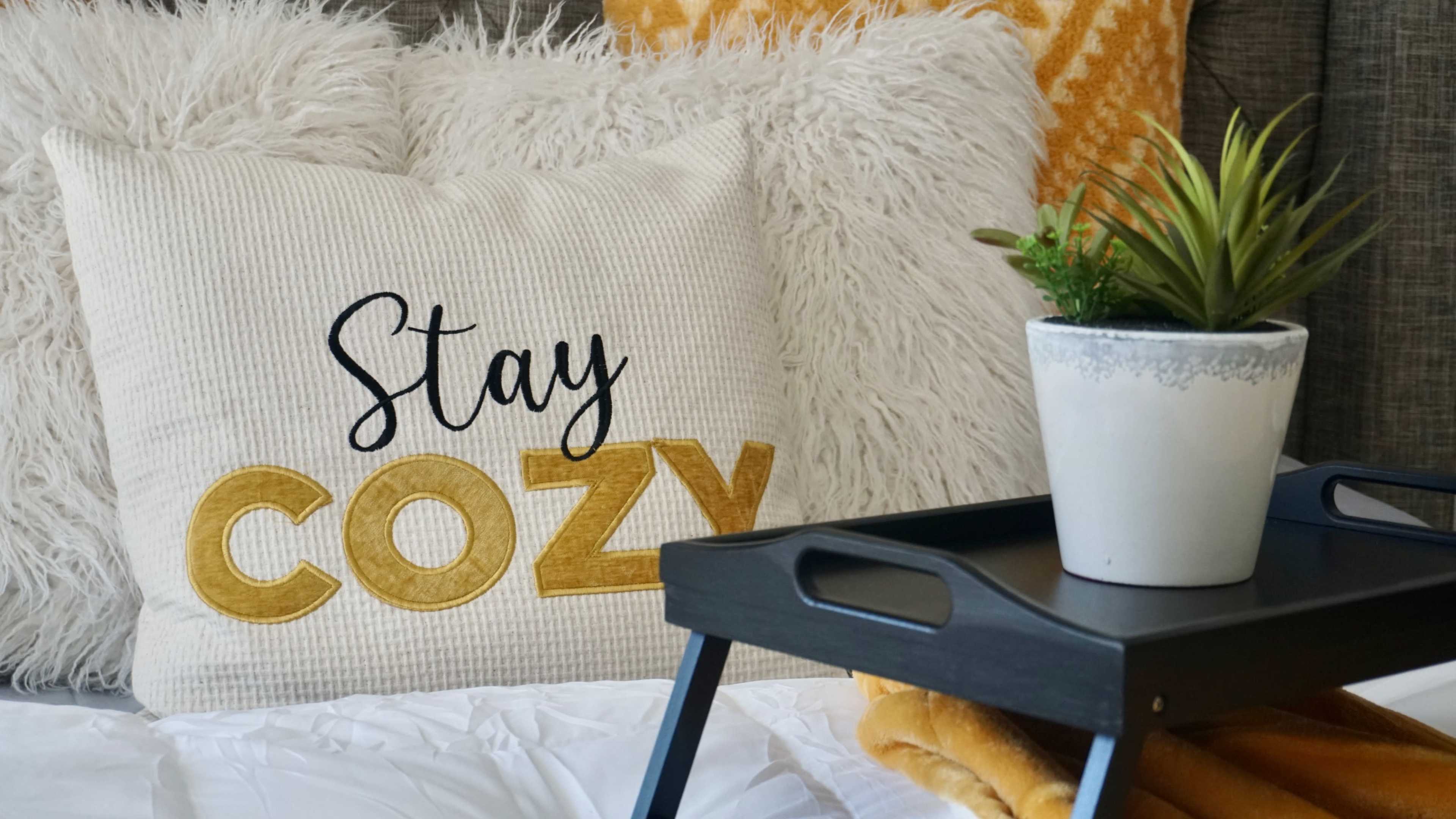 A decorative pillow that reads "Stay Cozy" on a bed with a small potted plant on a tray nearby.