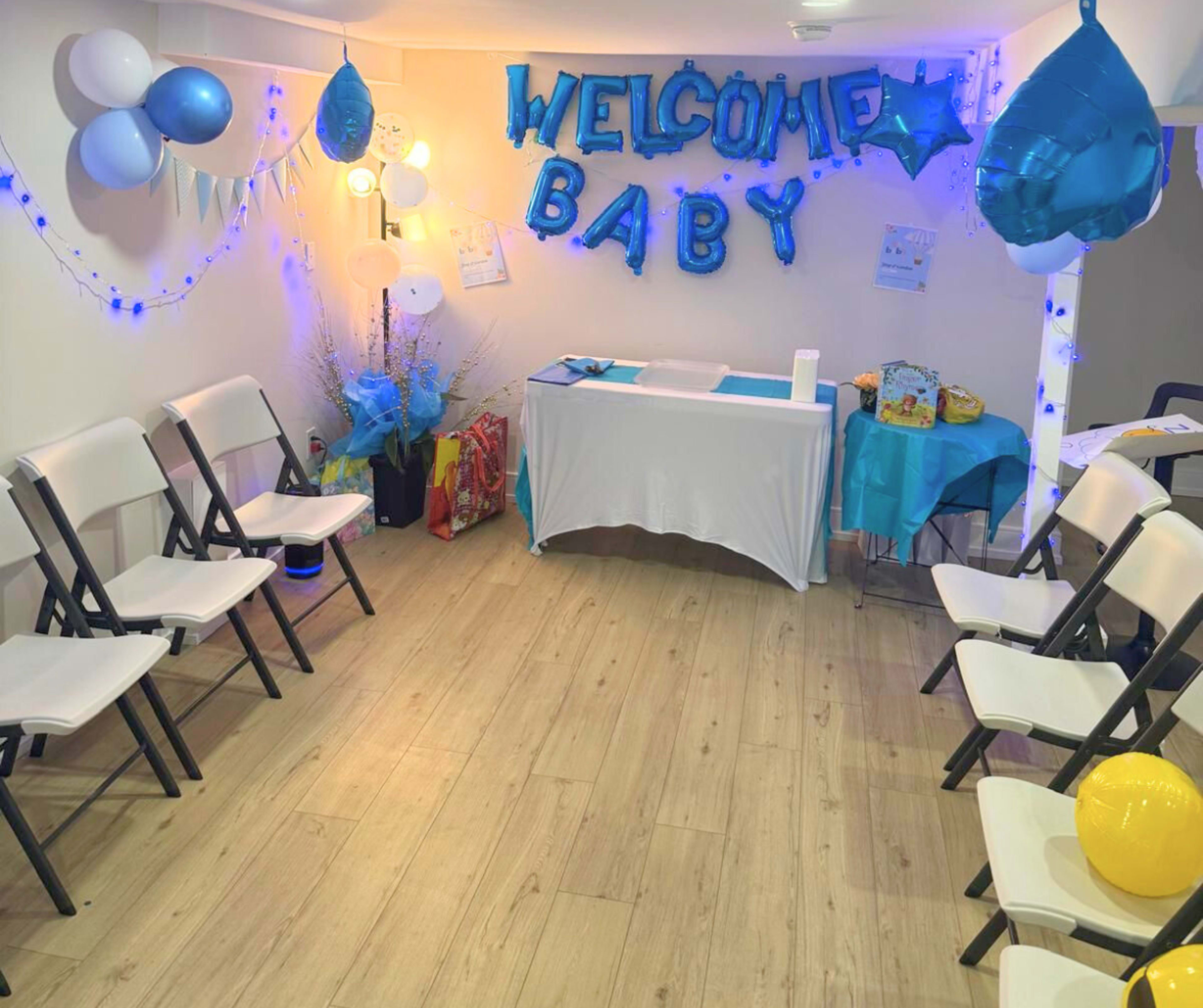 The image shows a decorated room set up for a baby shower, featuring blue balloons, a welcome sign, and seating arranged around a table.