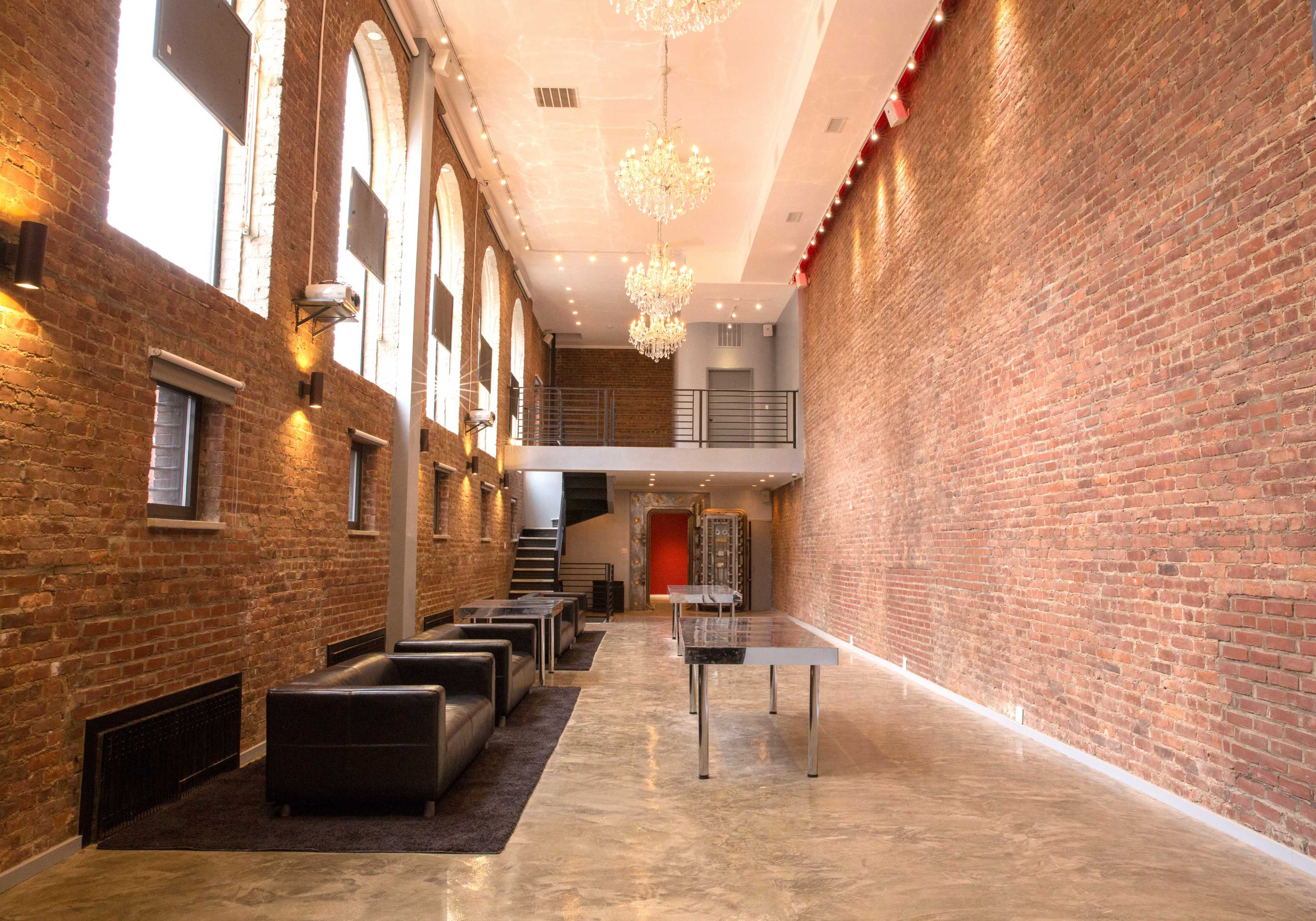 The image shows a spacious interior with exposed brick walls, large arched windows, modern chandeliers, and sleek furniture arranged along a polished concrete floor.