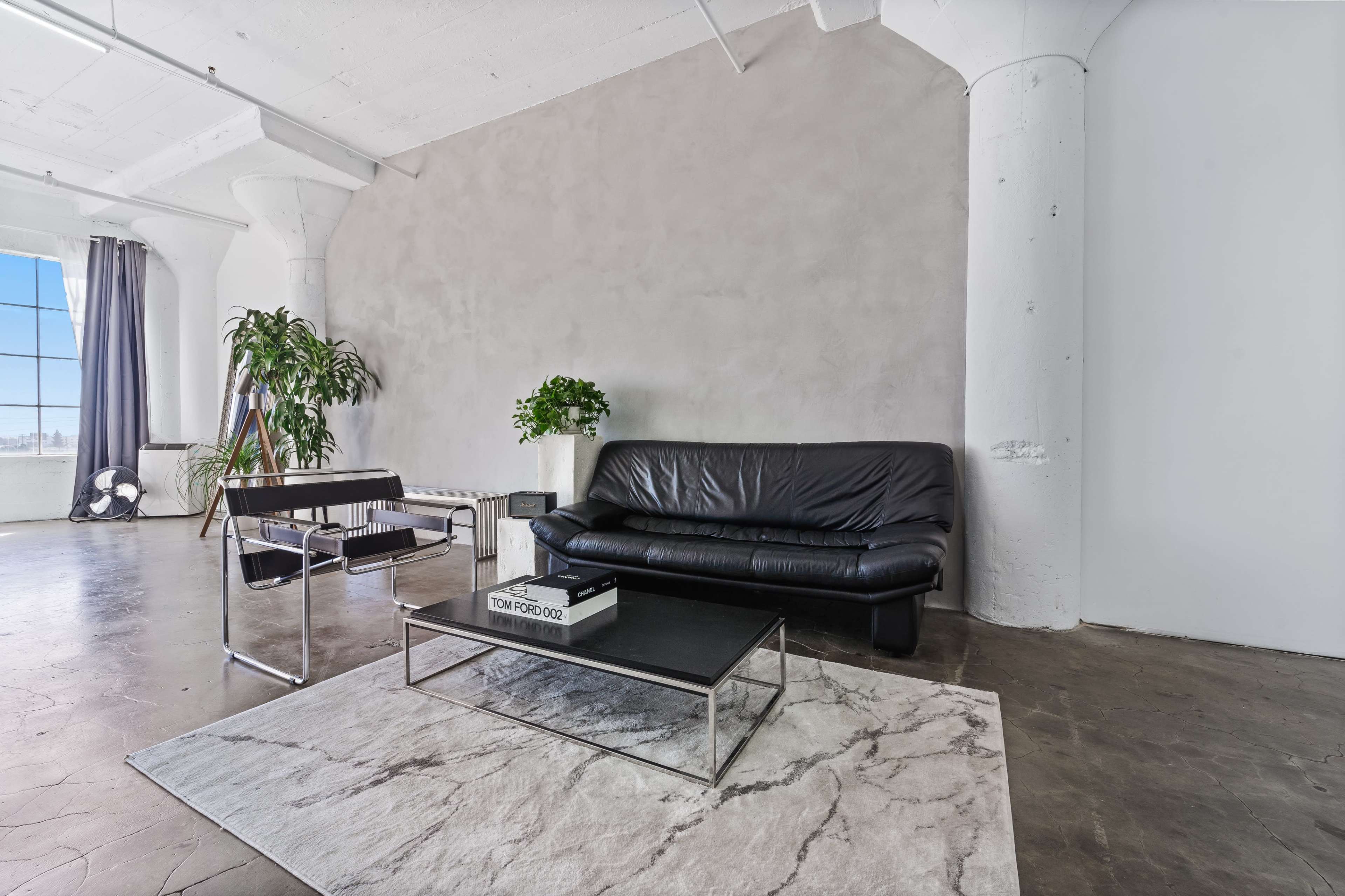 A modern living space featuring a black leather sofa, a glass coffee table, and a decorative plant beside a large window.