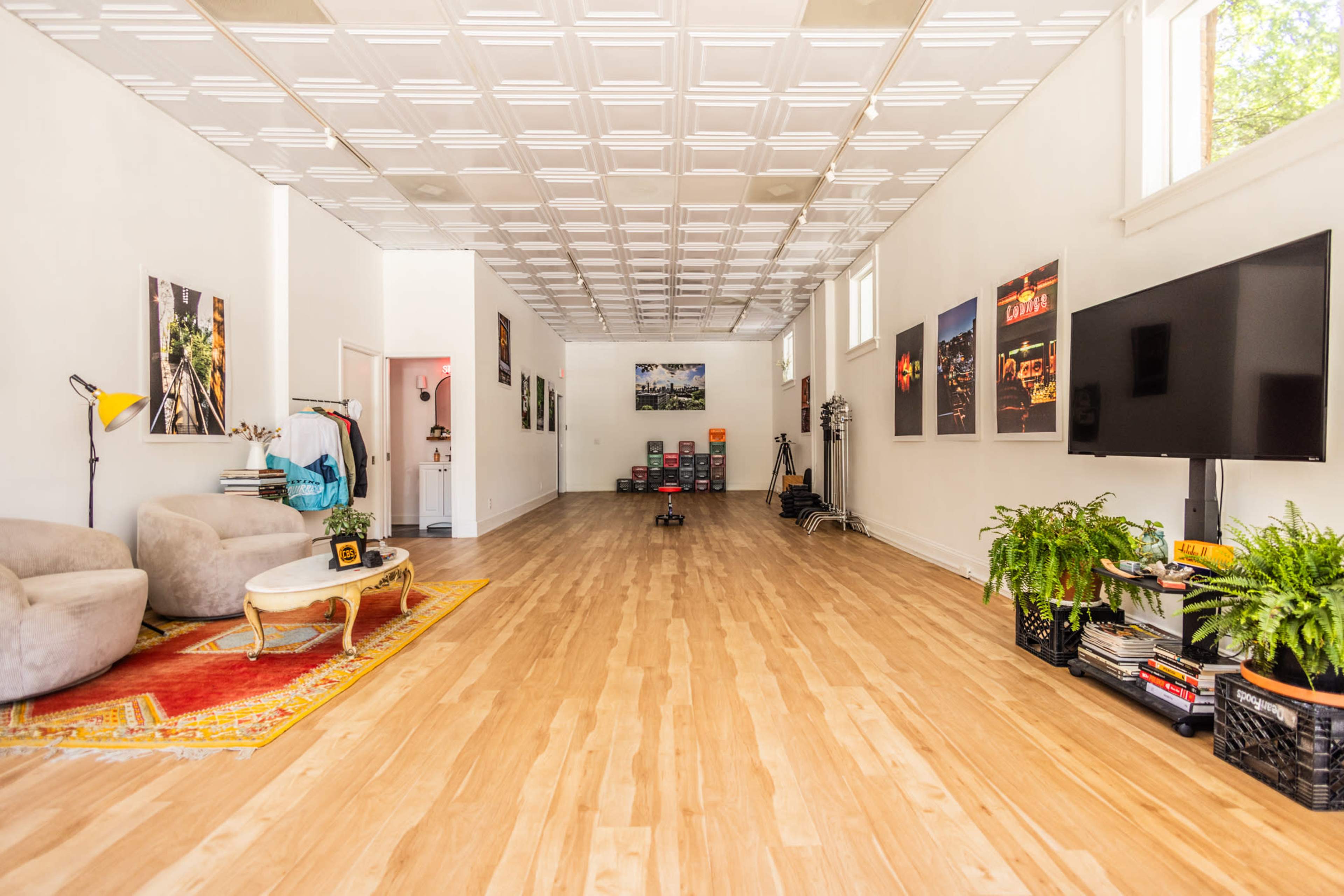 The image shows a spacious, bright room with light wood flooring, two gray armchairs, a coffee table, a television, and various framed artworks on the walls.