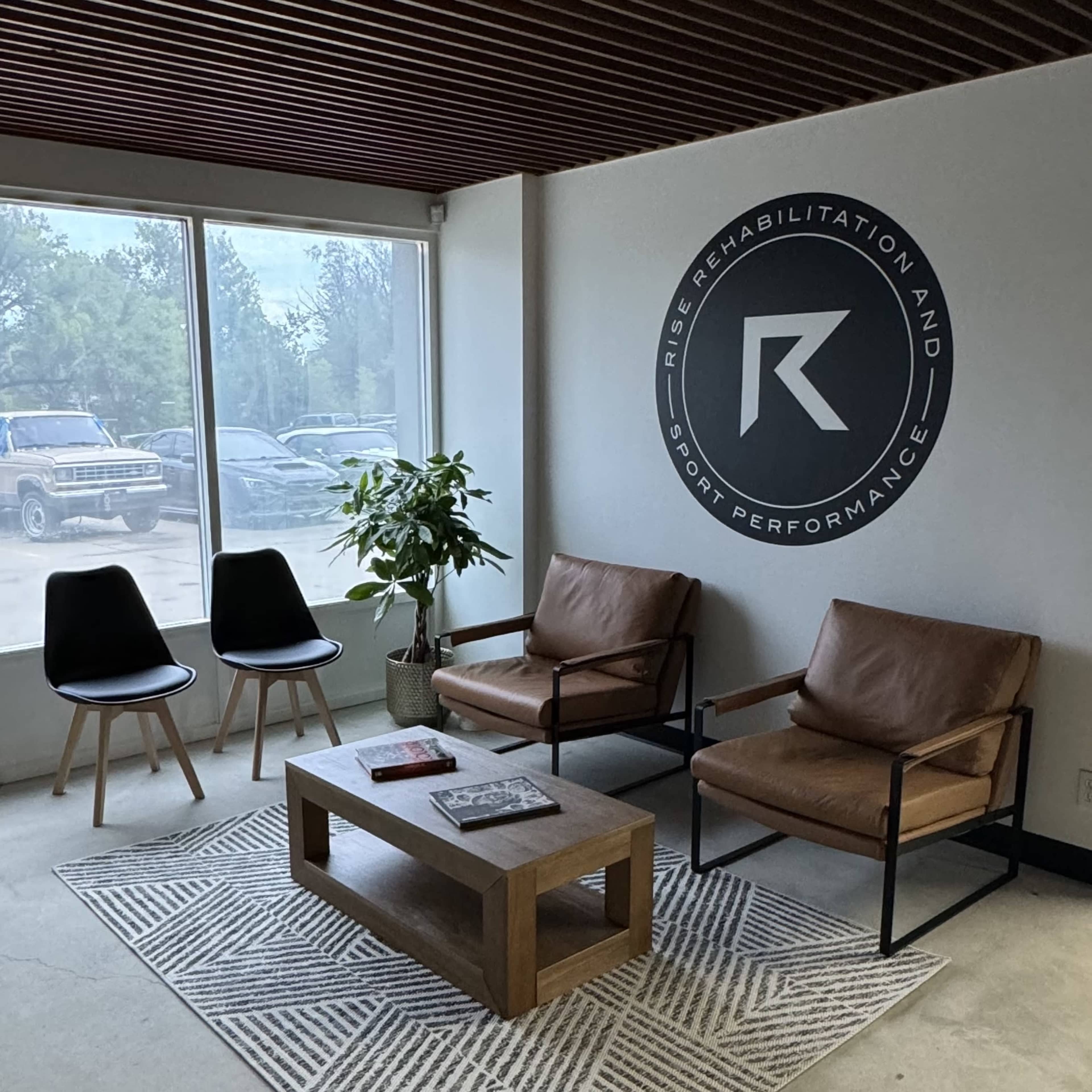 The image shows a waiting area with two brown chairs, a small wooden table, and a wall featuring a circular logo for "Rise Rehabilitation and Sport Performance."
