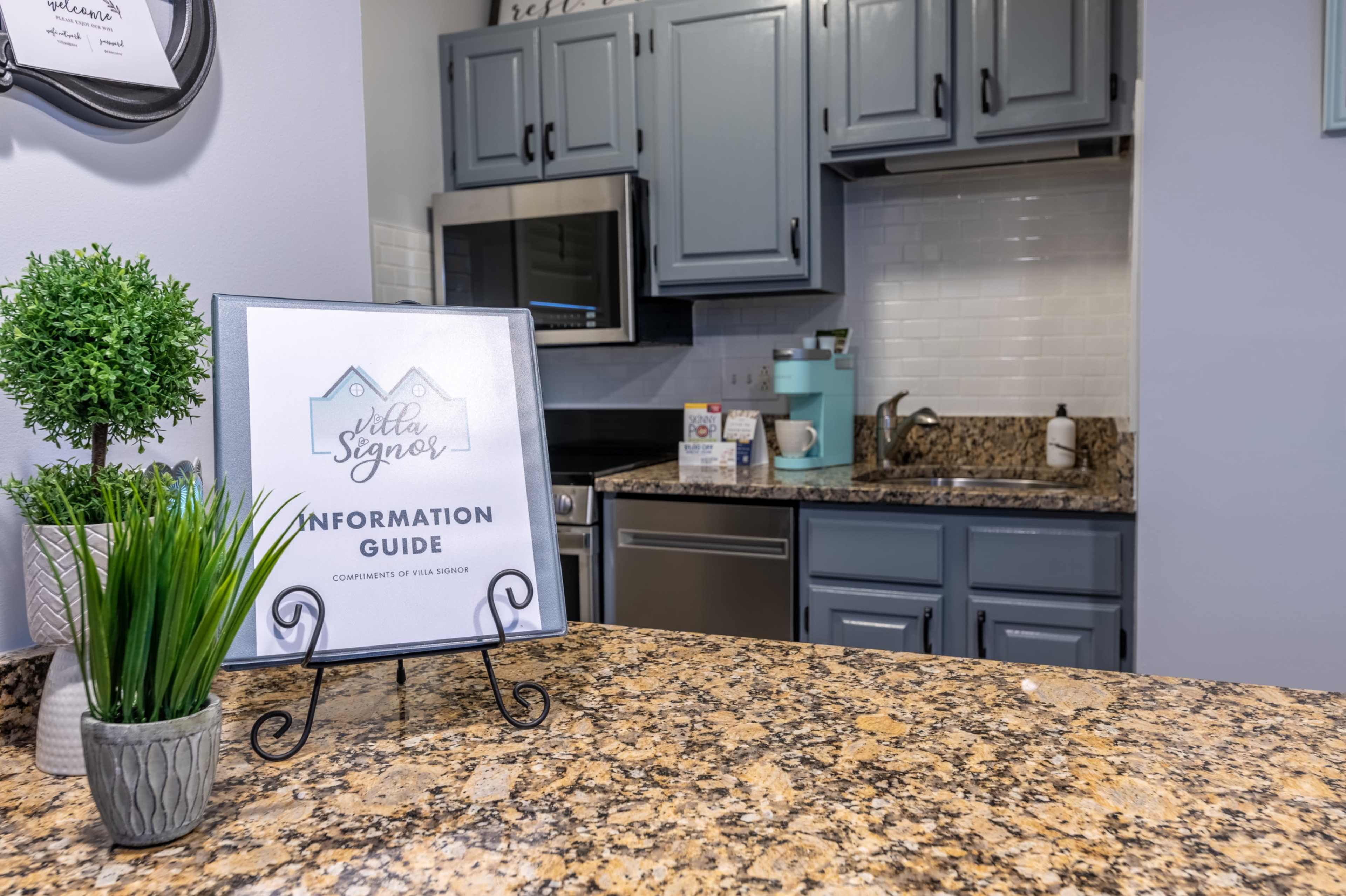 A kitchen with gray cabinets, a countertop made of speckled granite, and a sign labeled "Information Guide" on a stand in front of a small plant.