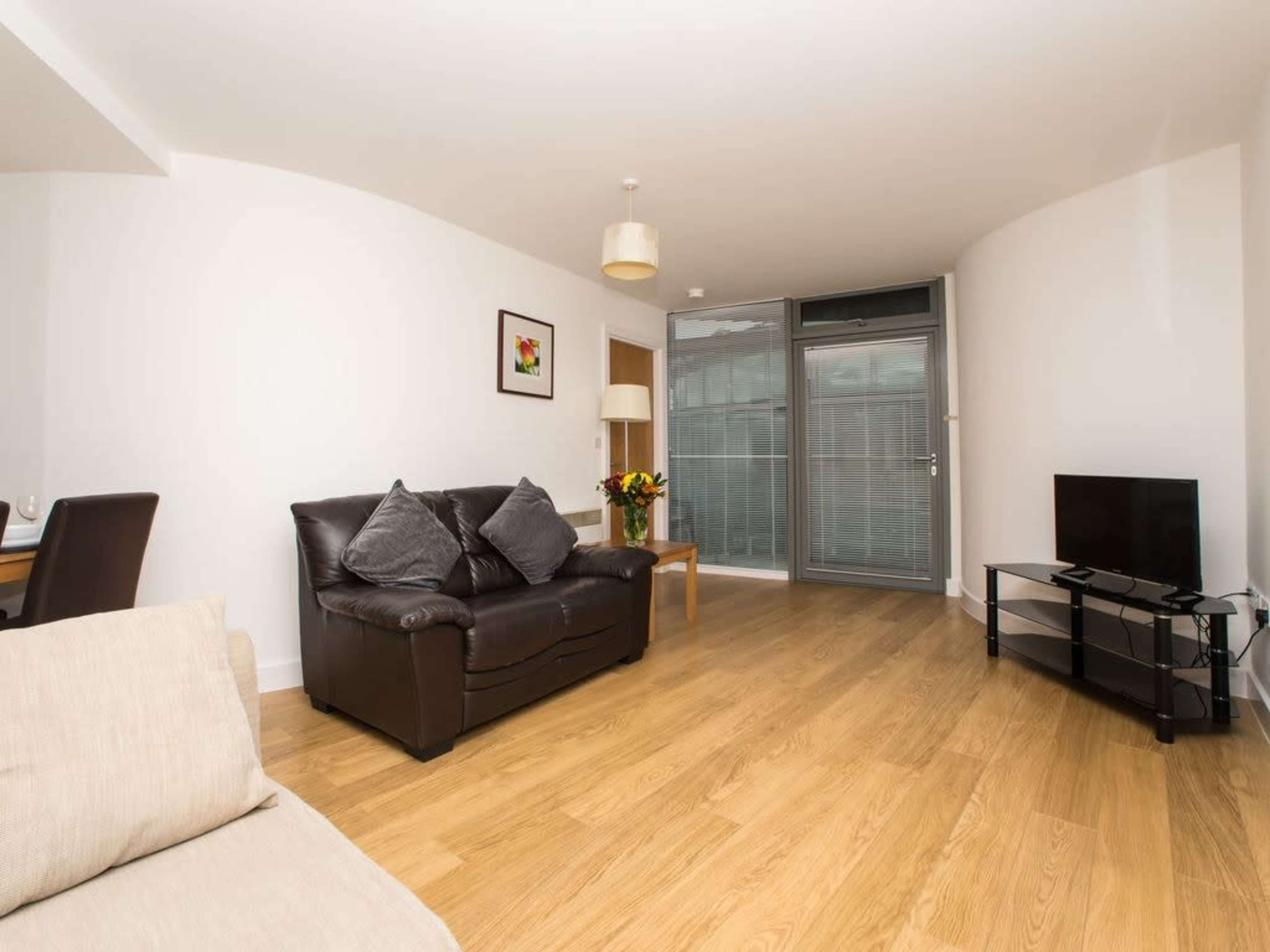 A living room with a brown leather sofa, a light-colored armchair, a television on a stand, and large windows with blinds, all situated on a wood-paneled floor.